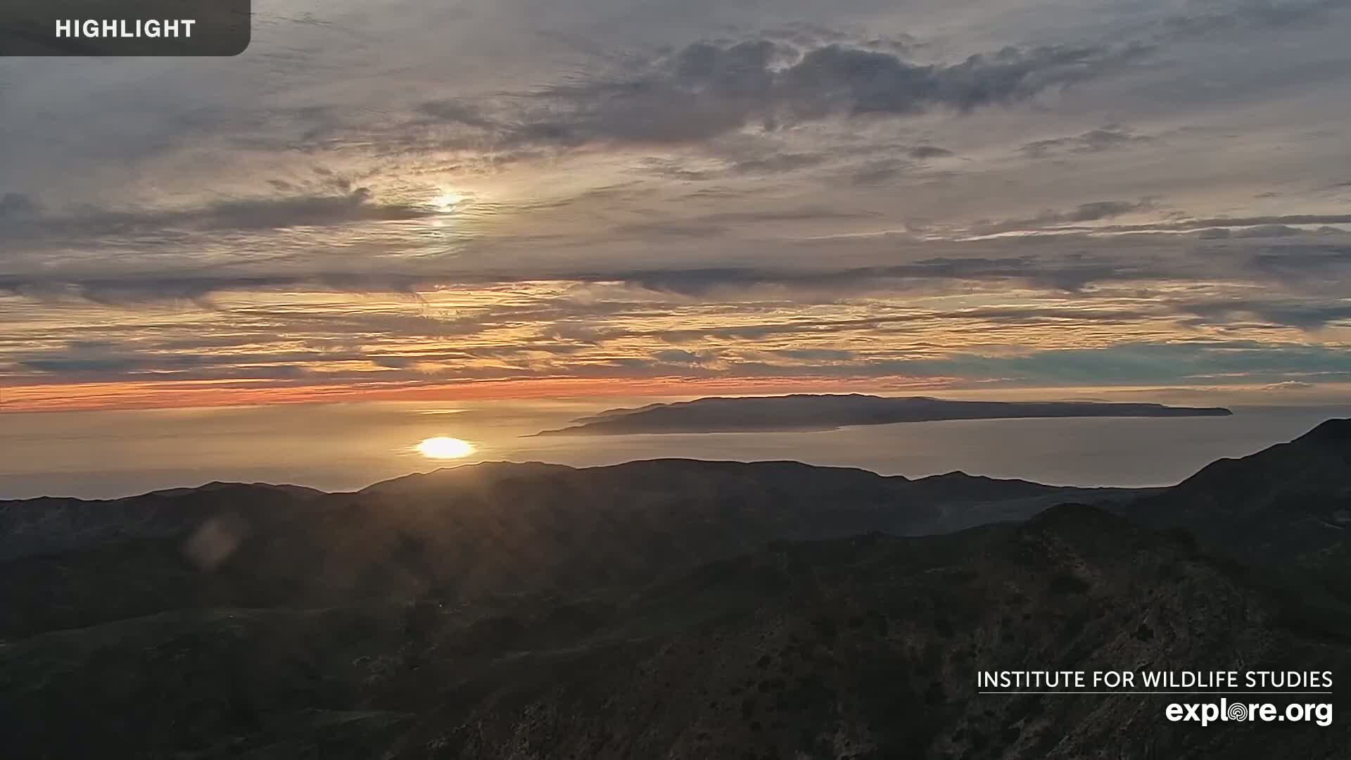 A sunset over a calm ocean and mountainous landscape, with a partly cloudy sky.
