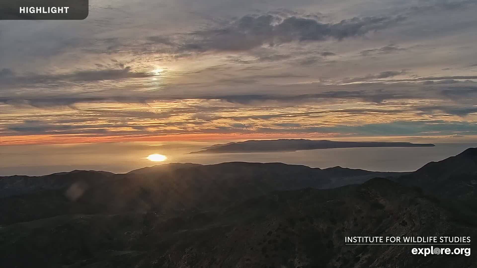 A sunset over a calm ocean and distant islands, viewed from a mountain range under a partly cloudy sky.