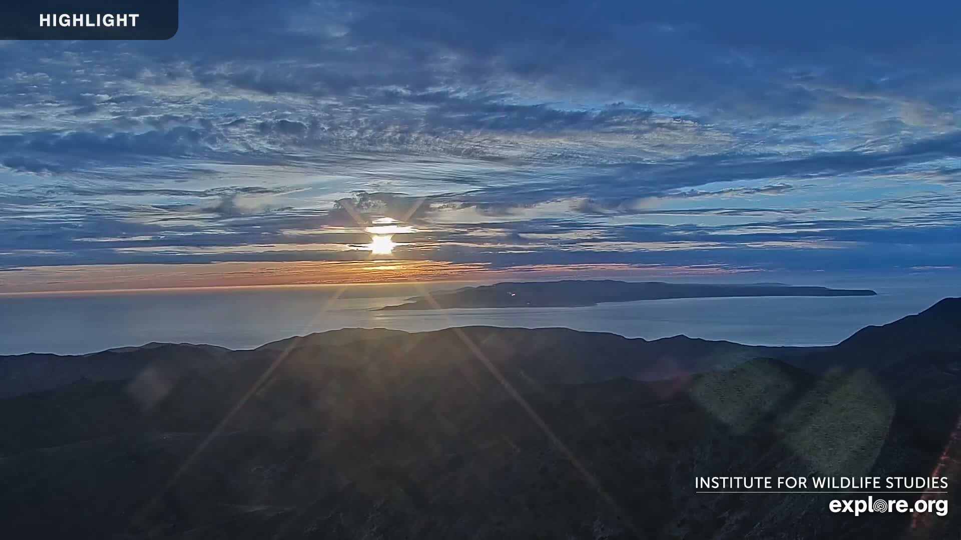 A sunset over a calm ocean and distant islands, viewed from a mountaintop under a partly cloudy sky.