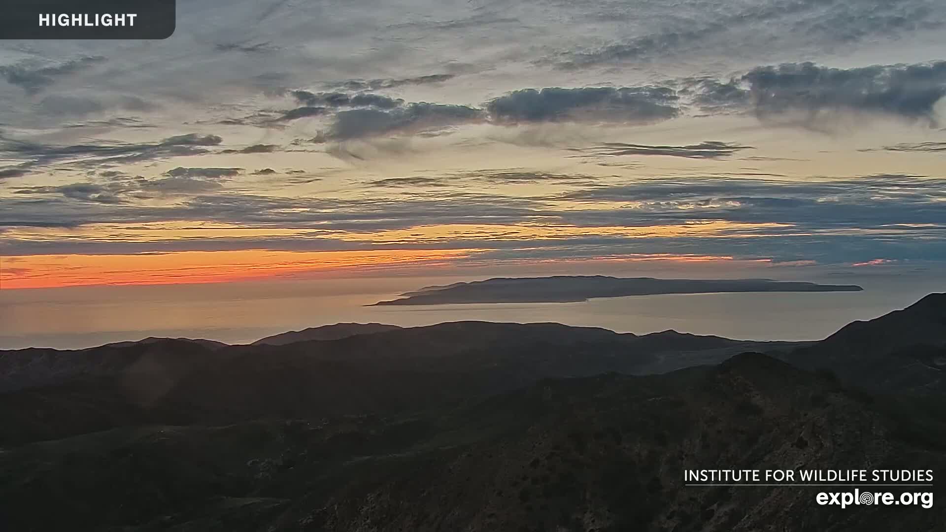 A sunset over a calm ocean with a distant island visible from a mountaintop.