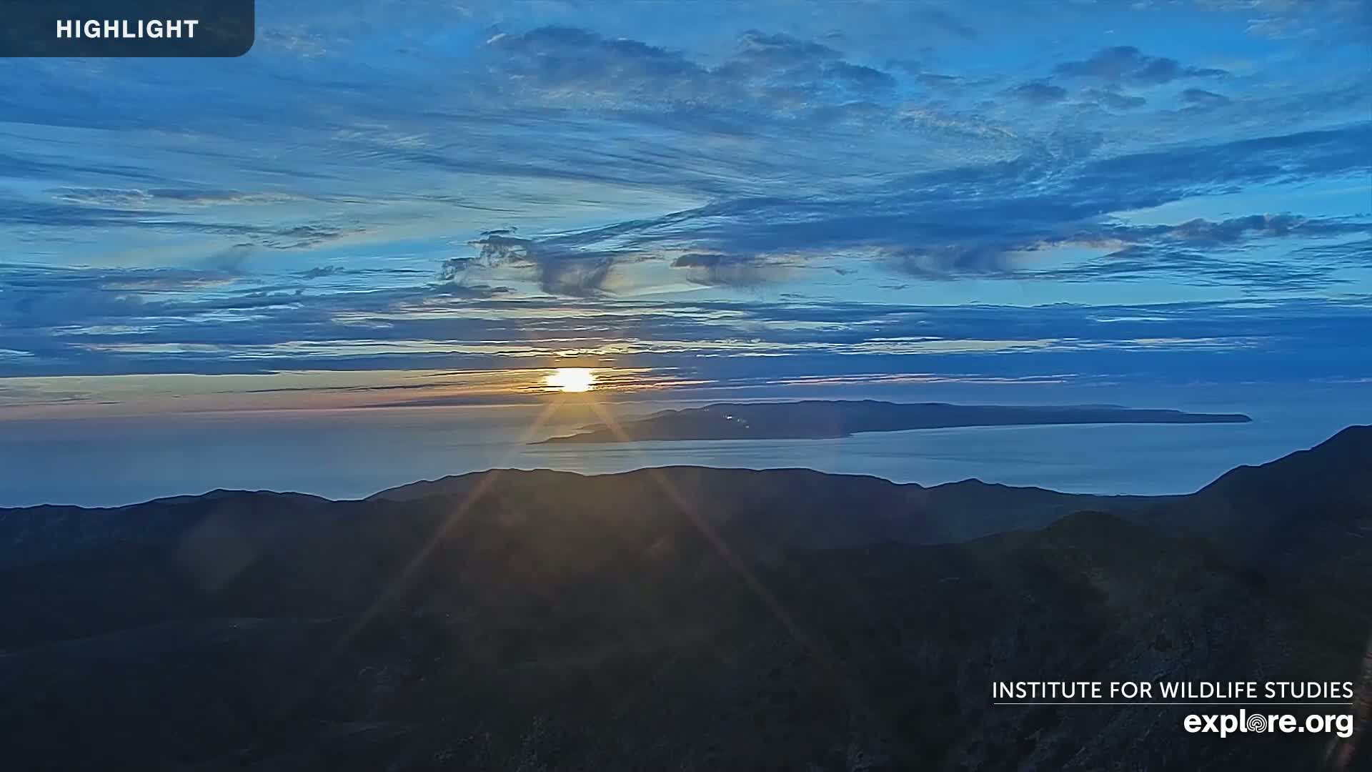A sunrise over a calm ocean and distant islands, viewed from a mountaintop under a mostly clear, blue sky with scattered clouds.