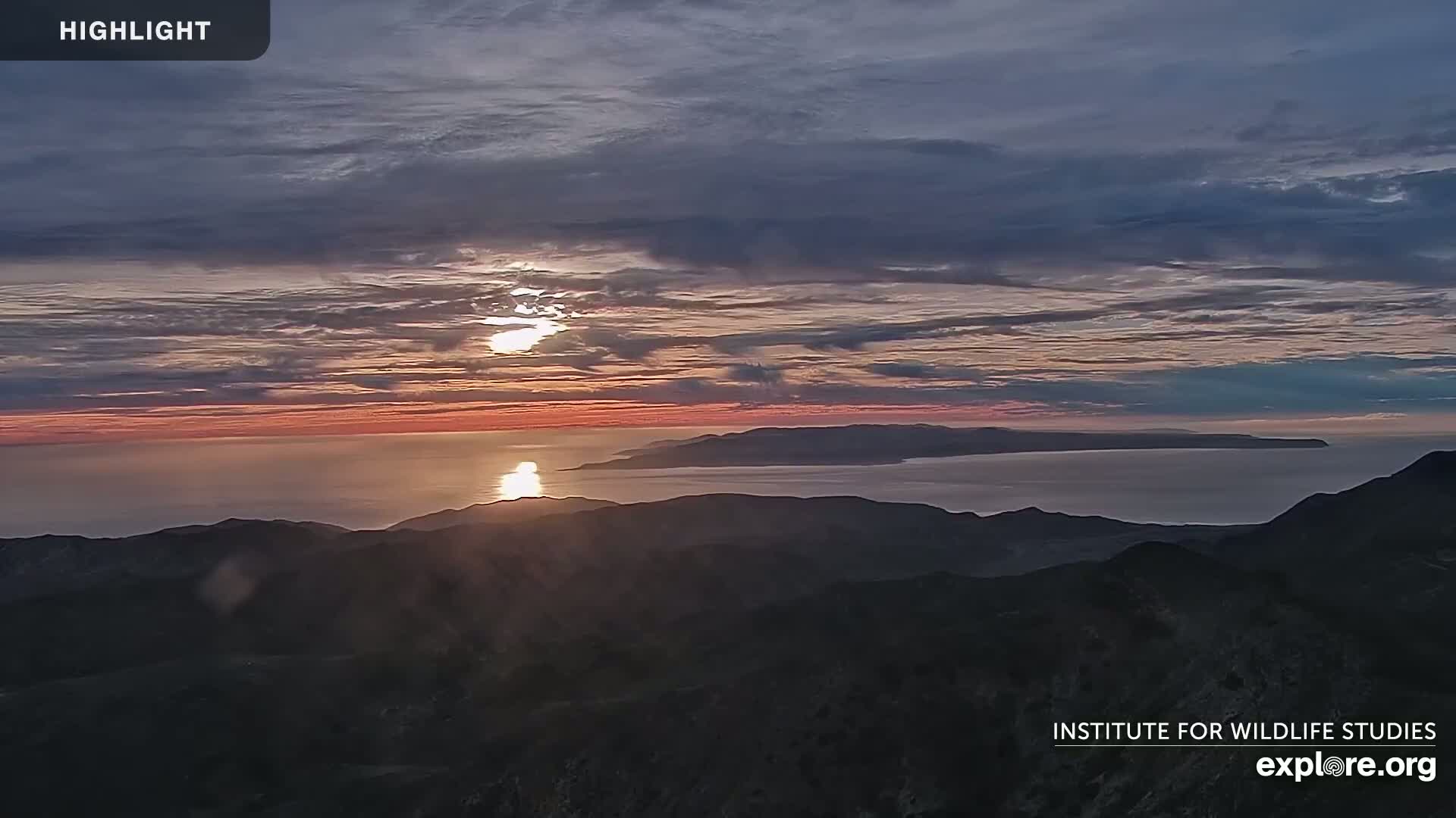 A sunset over a calm ocean, with a distant island visible and mountains in the foreground under a partly cloudy sky.