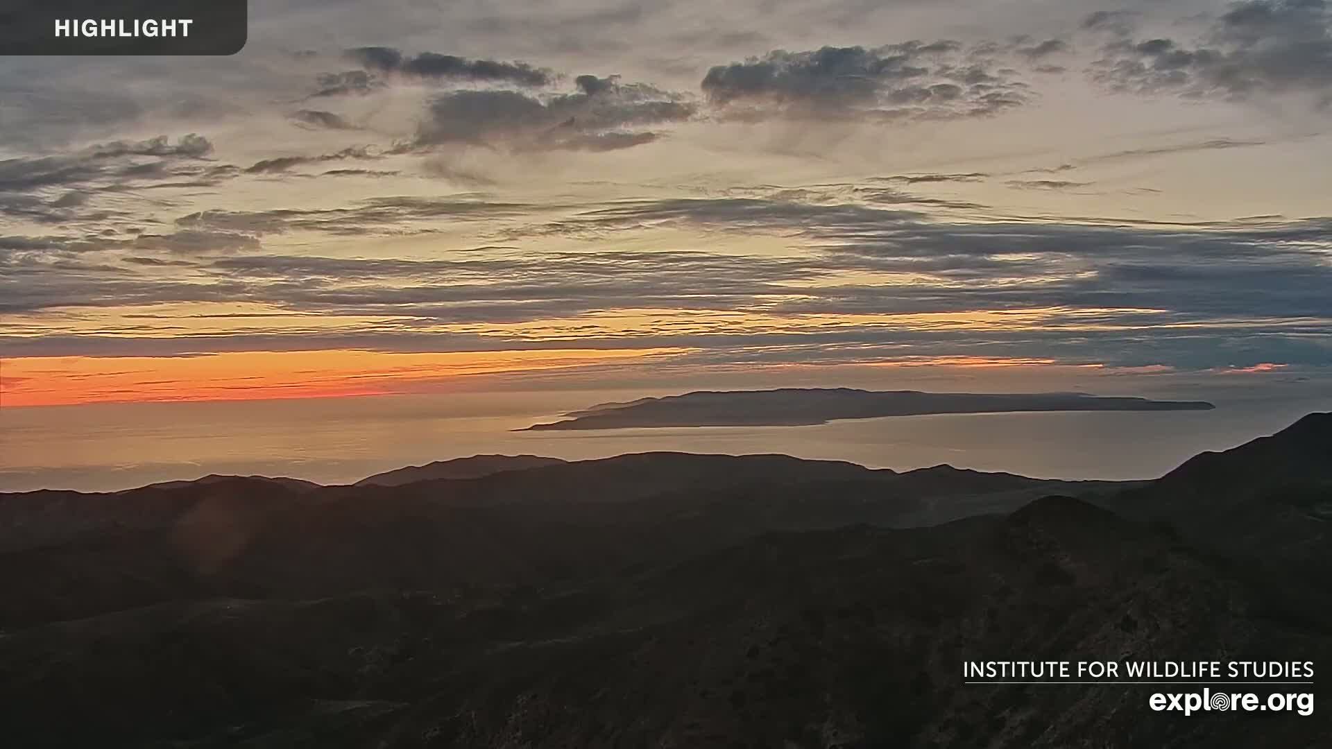 A sunrise paints the sky with orange and pink hues over a calm ocean separating dark mountain silhouettes.