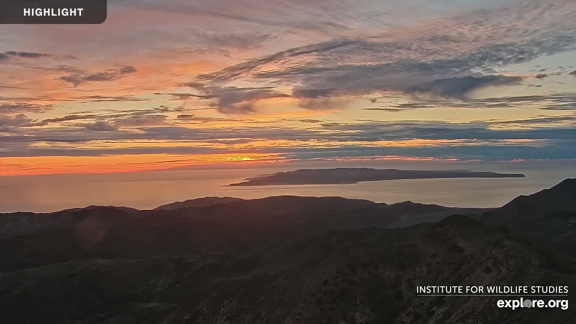 A sunset over a calm ocean and mountainous landscape, with a sliver of land visible in the distance.