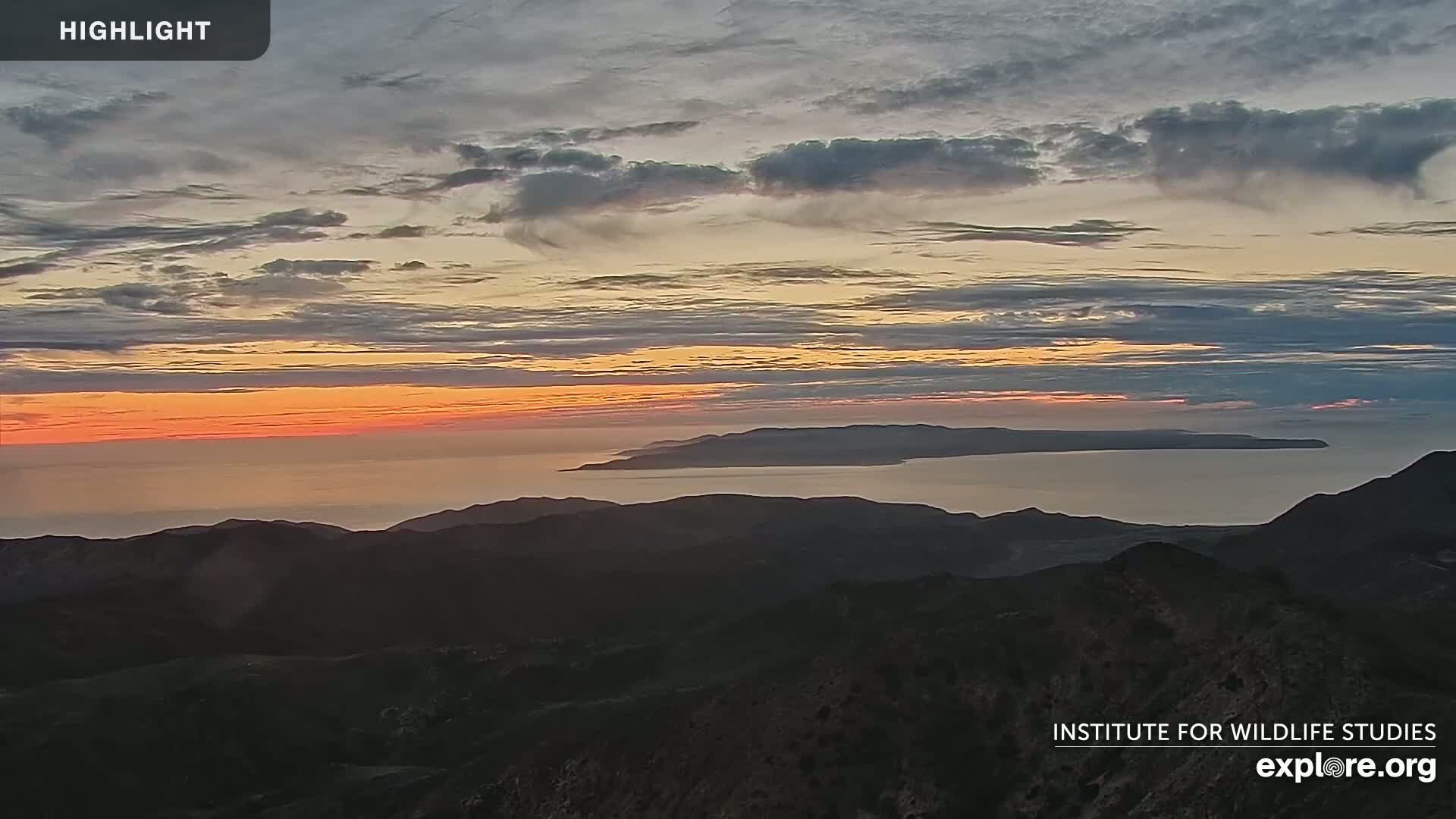 A sunset over a calm ocean, with an island visible in the distance and dark mountains in the foreground under a partly cloudy sky.