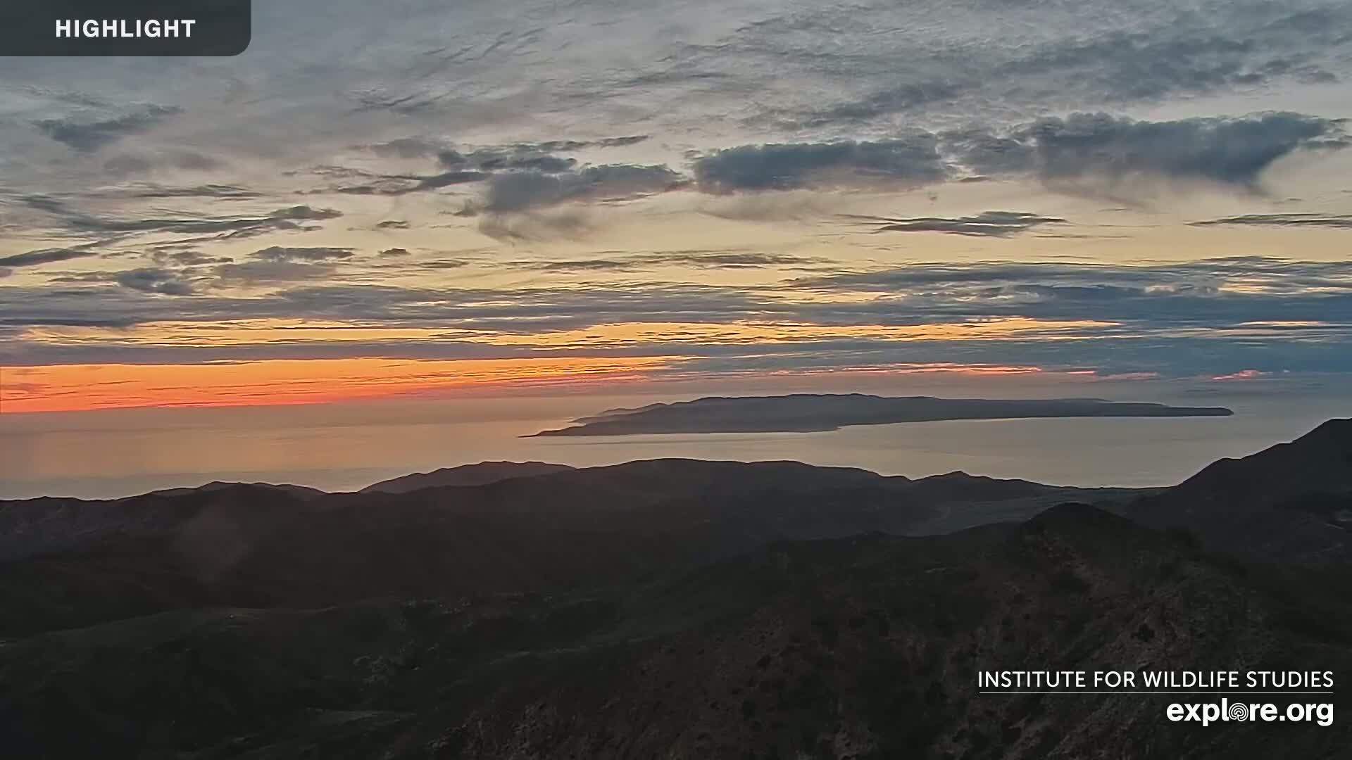 A sunset over a calm ocean and distant islands, viewed from a mountaintop under a partly cloudy sky.