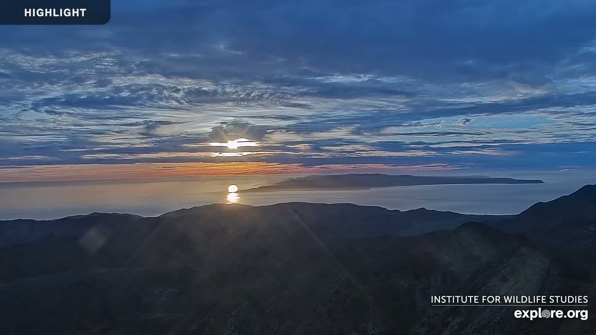 A sunset over a calm ocean and distant islands is viewed from a mountaintop under a partly cloudy sky.