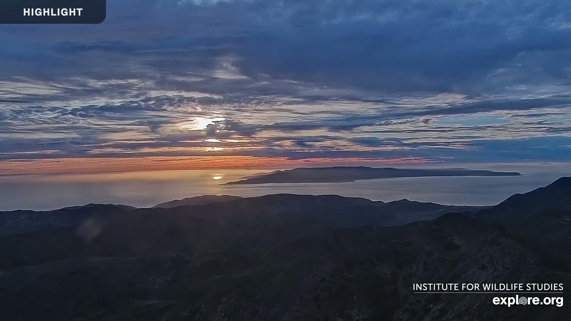 A sunset over a calm ocean and distant islands is viewed from a mountaintop under a partly cloudy sky.