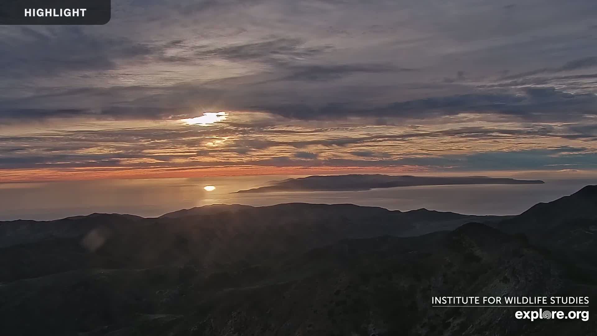 A sunset over a calm ocean and distant islands, viewed from a mountaintop.