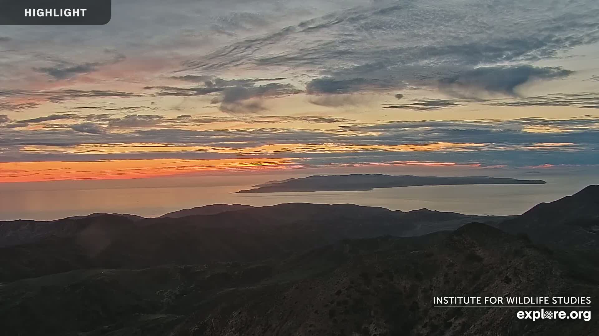 A sunset over a calm ocean and distant islands is viewed from a mountain range.