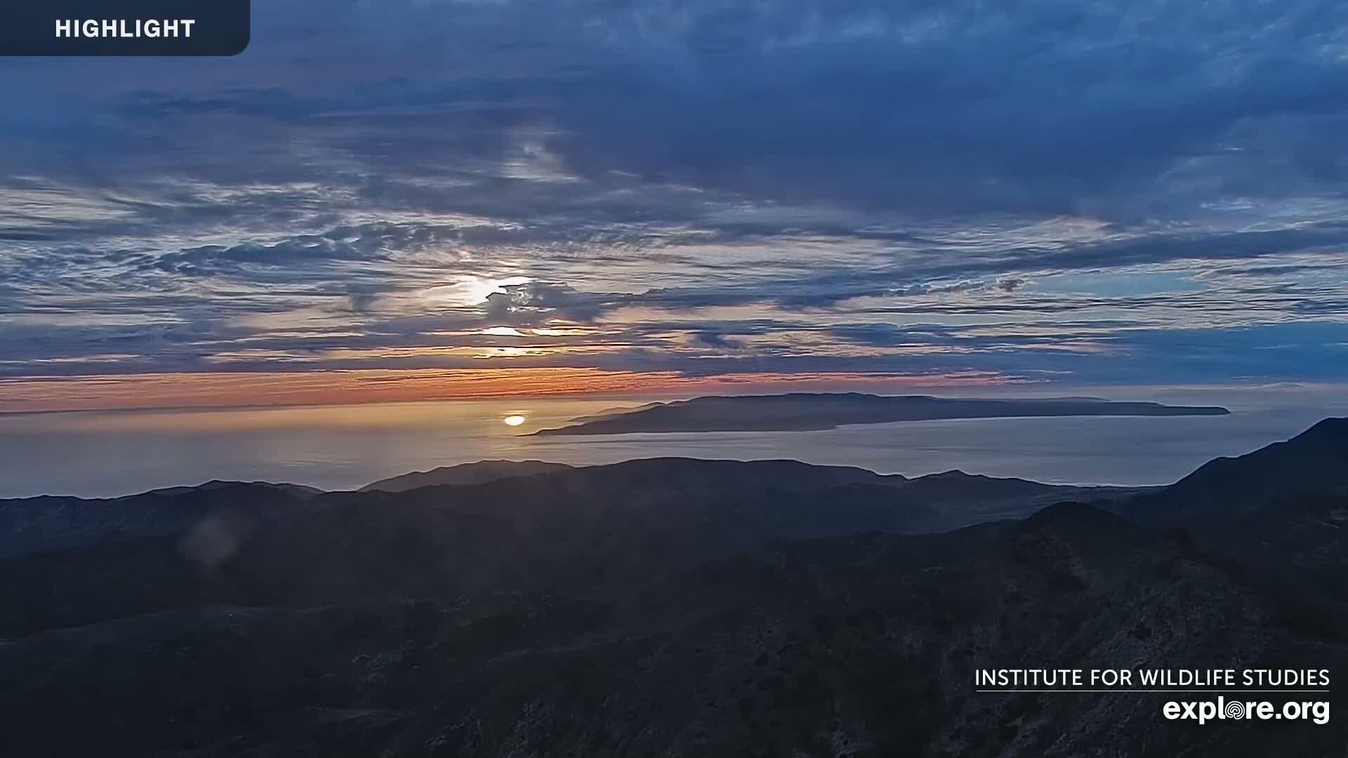 A sunset over a calm ocean and distant island is viewed from a mountaintop under a partly cloudy sky.