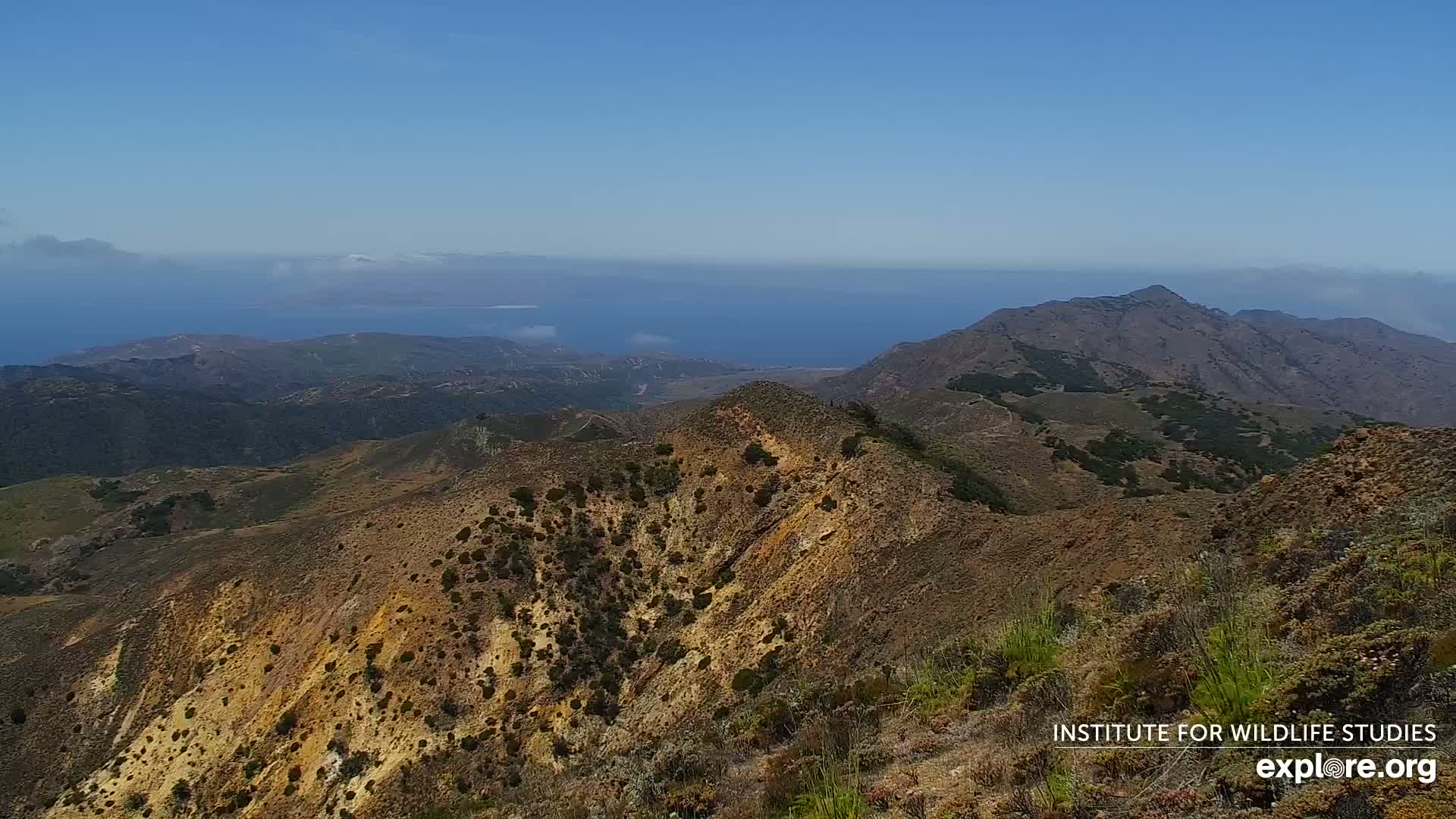A high-angle view shows a range of arid, brown and tan mountains overlooking a distant ocean on a clear, sunny day.