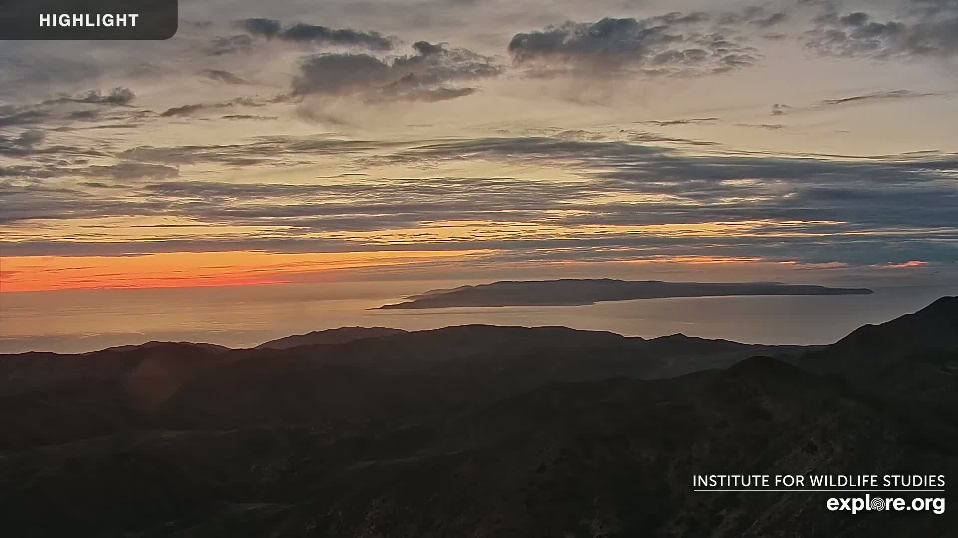 A sunset over a calm ocean, with islands visible in the distance and dark mountain ranges in the foreground.