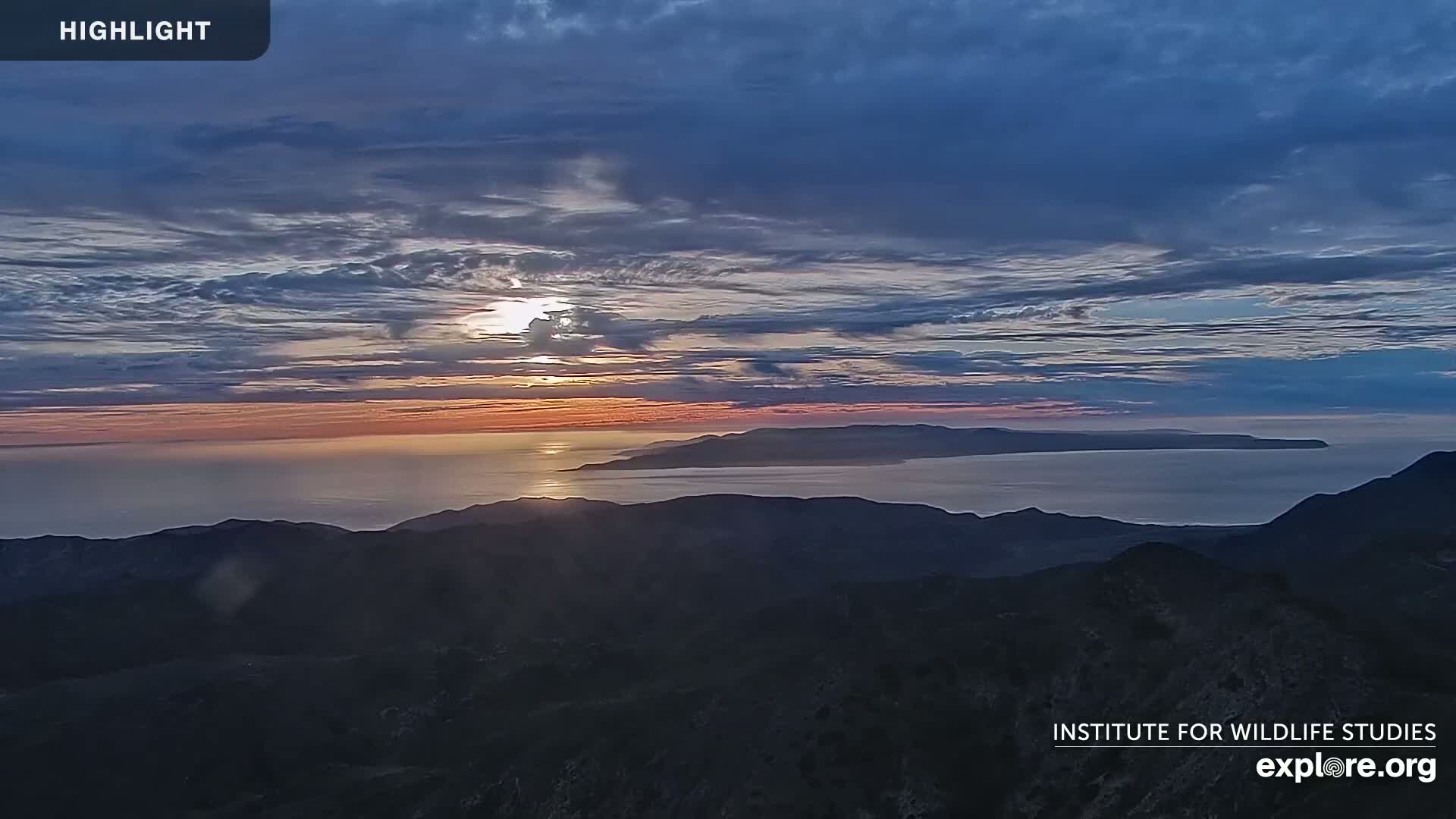 A sunset over a calm ocean, with islands visible in the distance and dark mountain ranges in the foreground.