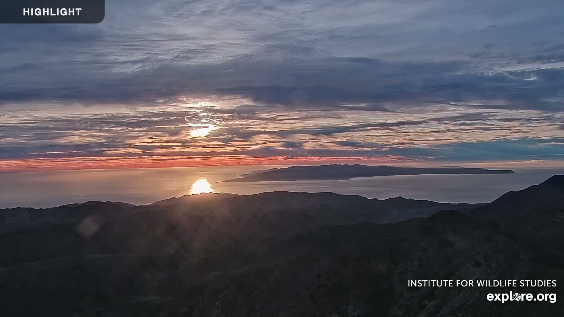 A sunrise over a calm ocean, illuminating a distant island and mountain range below a partly cloudy sky.