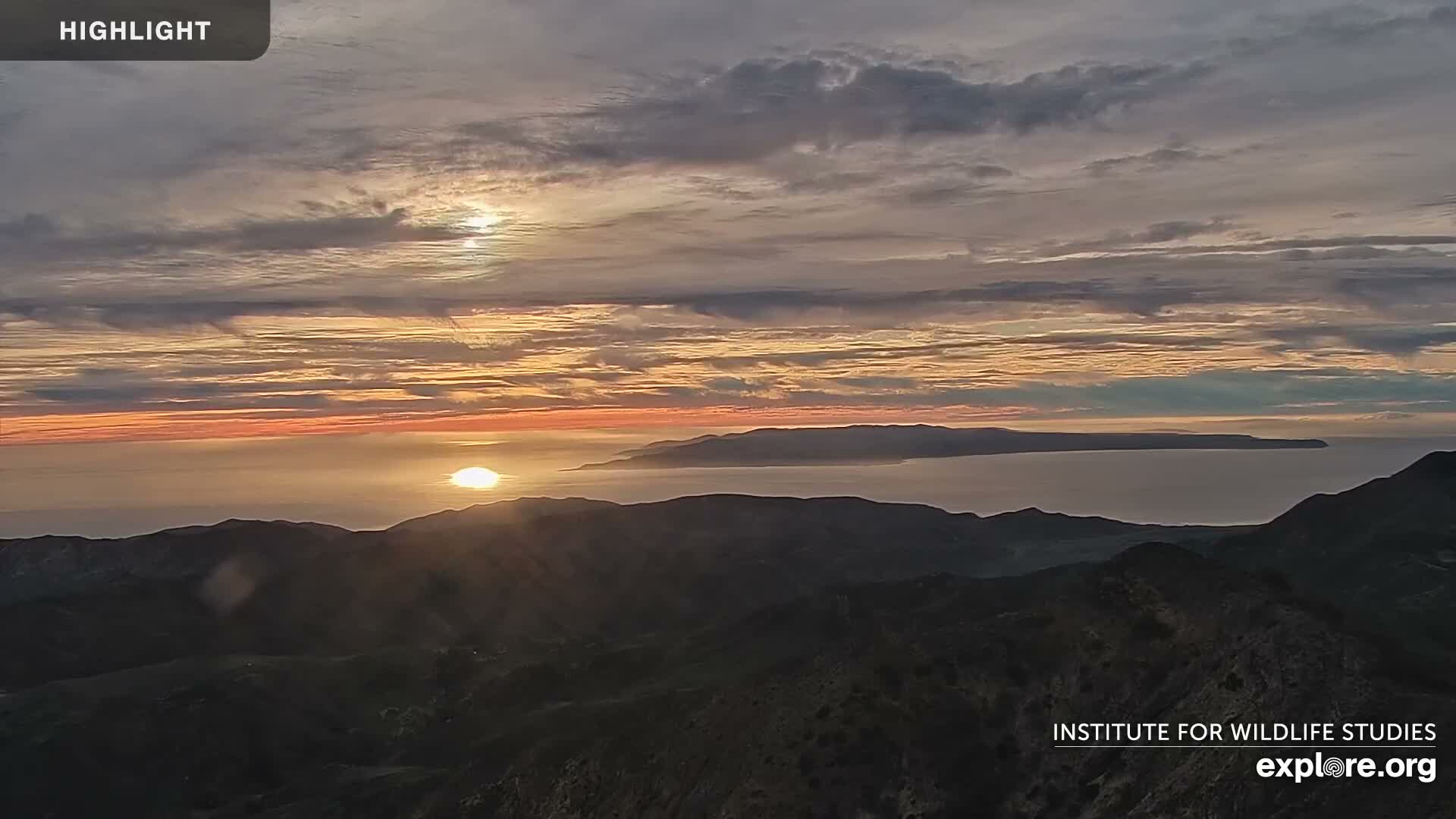 A sunrise over a calm ocean, illuminating a distant island and mountain range below a partly cloudy sky.