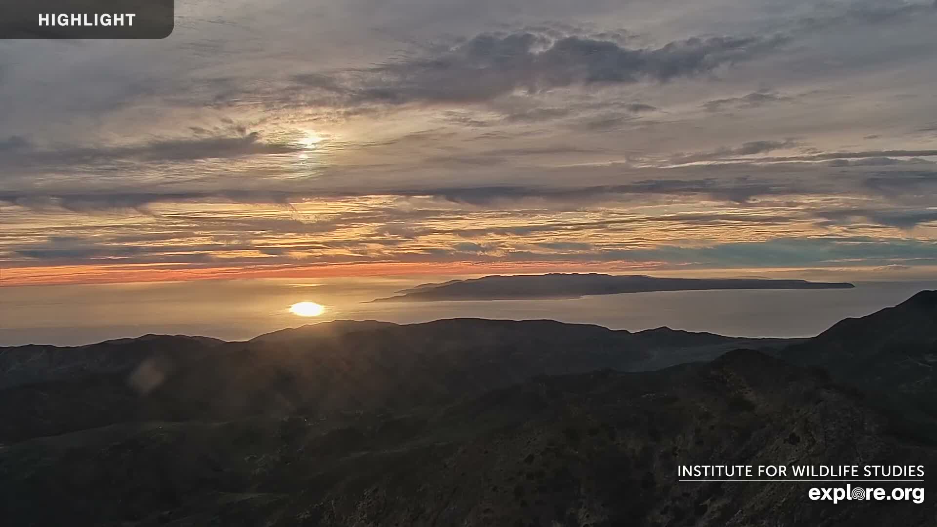 A cloudy sky with warm orange and gray tones frames a sunset or sunrise over the ocean, with the sun's light breaking through clouds and reflecting on the water, illuminating a dark mountainous foreground and a long, distant island.