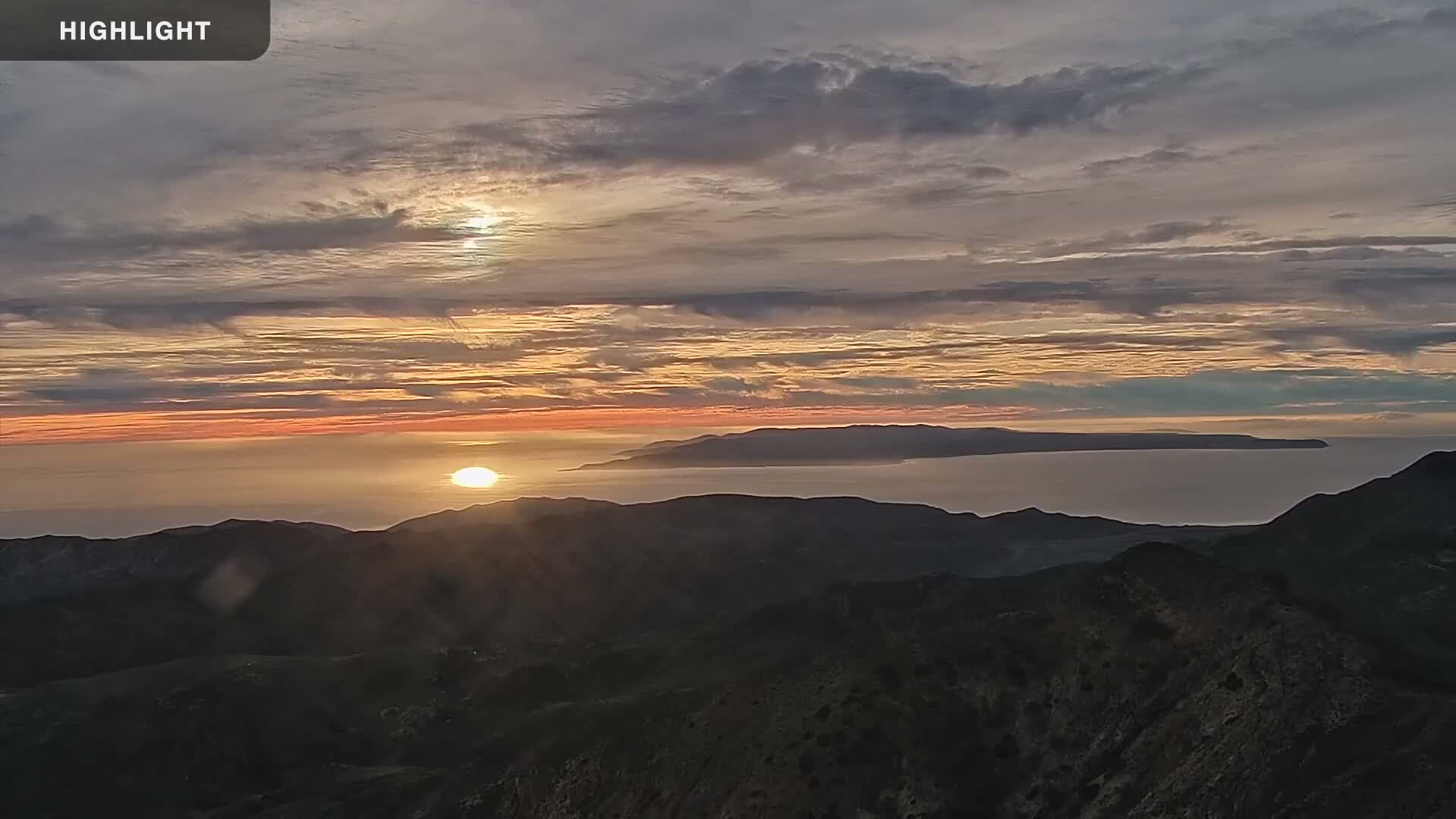 A serene sunset illuminates a hazy ocean with a long distant island, and dark, rolling hills in the foreground, under a partly cloudy sky.