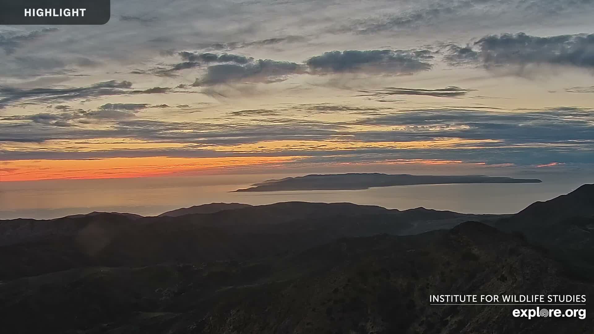 An expansive panoramic view at sunset reveals a calm ocean separating dark rolling hills in the foreground from a long, distant island, all beneath a sky filled with layered clouds painted in vivid oranges, yellows, and grays.