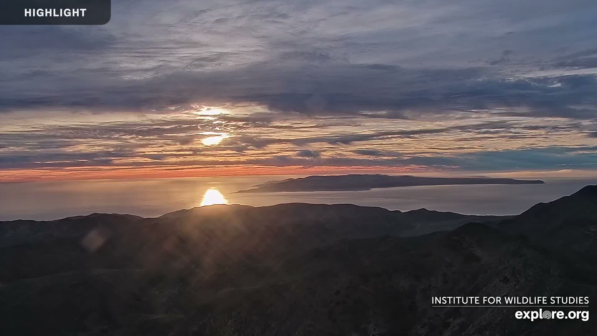 A bright sunset illuminates a partly cloudy sky and reflects on the ocean, framed by dark mountain ridges in the foreground and a long, low island silhouetted on the distant horizon.