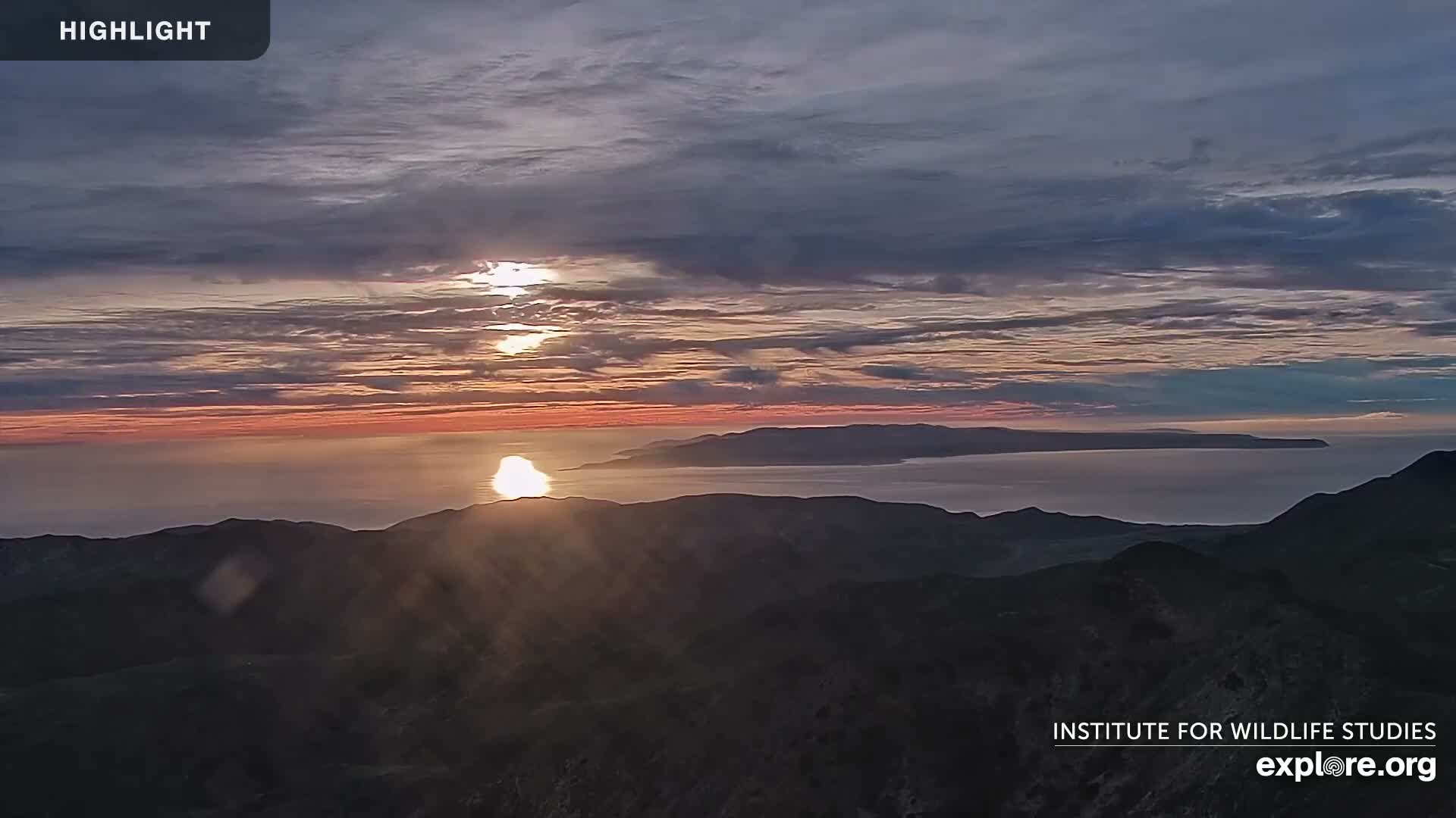 A colorful sunrise or sunset illuminates a partly cloudy sky with warm hues of orange and pink, casting a brilliant reflection on the calm ocean waters, with dark mountainous terrain in the foreground and a distant island silhouette under fair conditions.