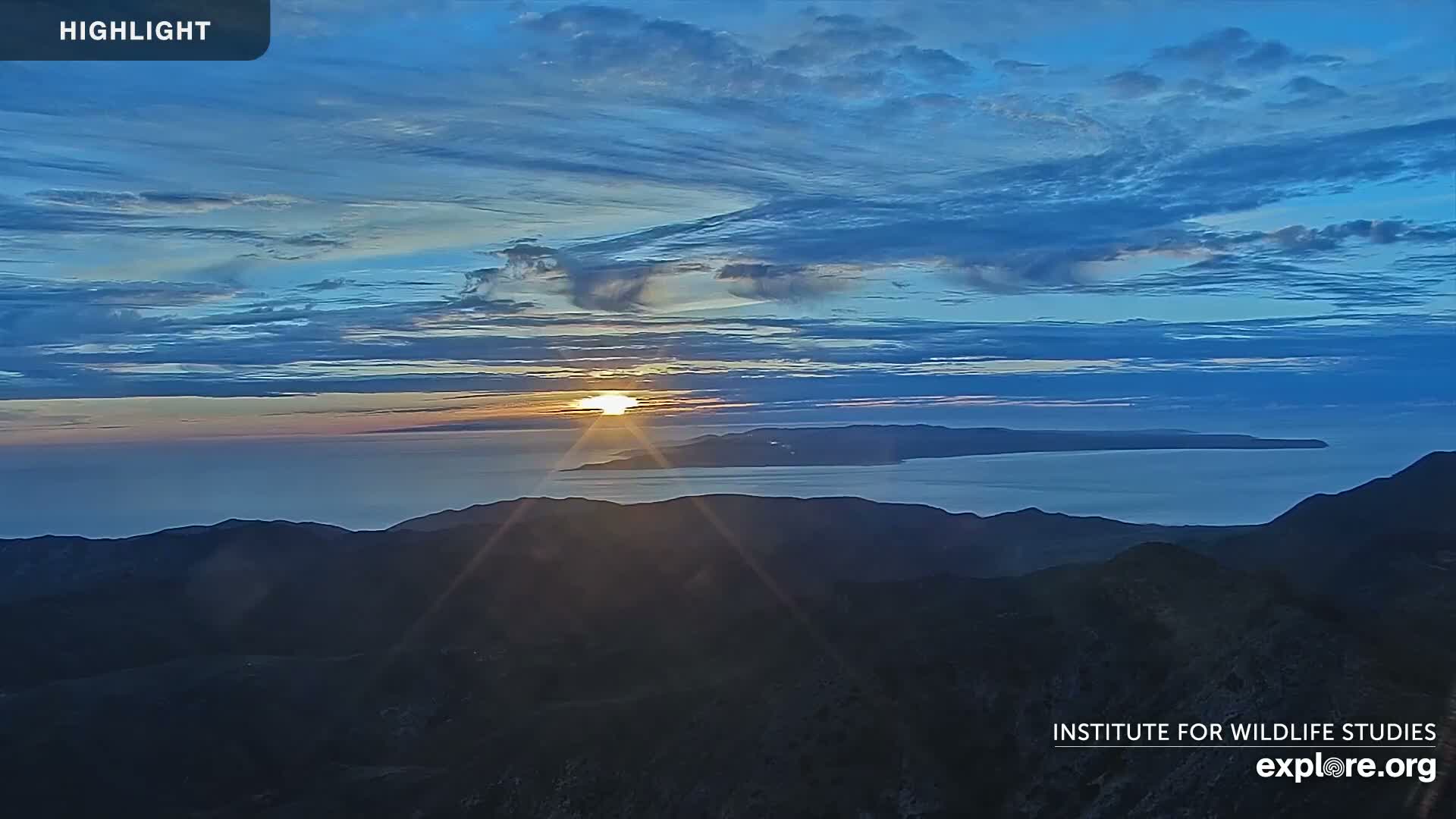 A colorful sunrise or sunset illuminates a partly cloudy sky with warm hues of orange and pink, casting a brilliant reflection on the calm ocean waters, with dark mountainous terrain in the foreground and a distant island silhouette under fair conditions.