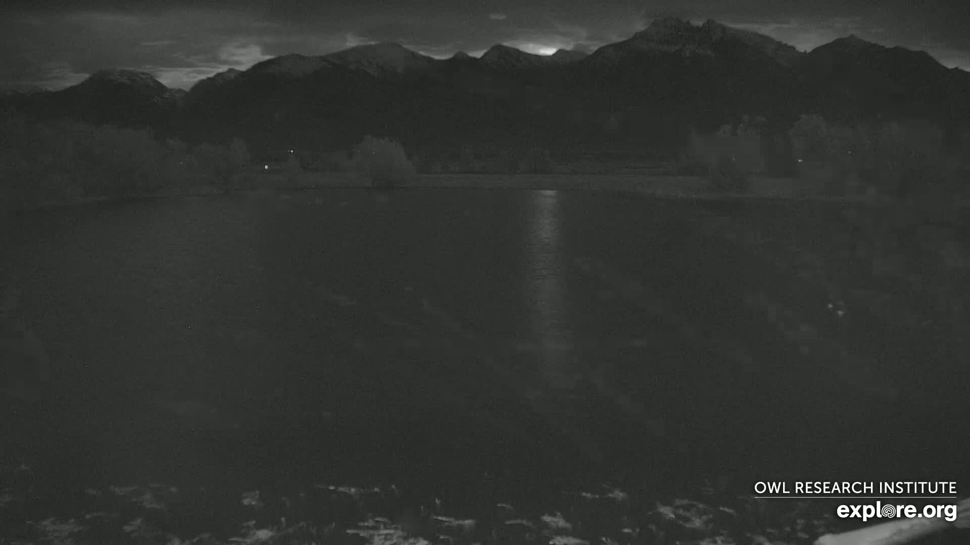 A dark, still body of water reflects the light from a mostly cloudy evening sky against a backdrop of silhouetted mountains.