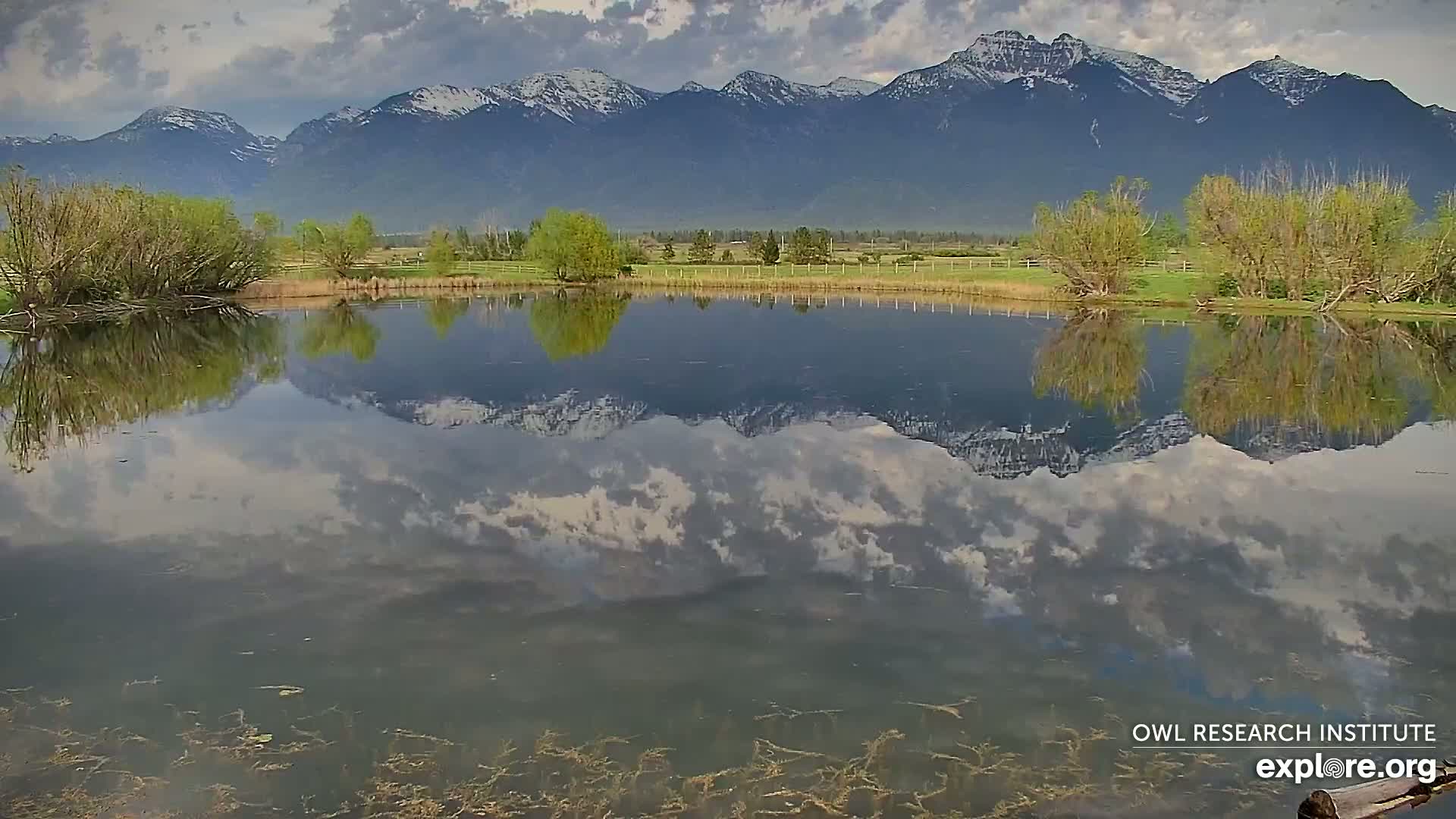 A calm pond reflects a partly cloudy sky and snow-capped mountains under a partly cloudy sky.