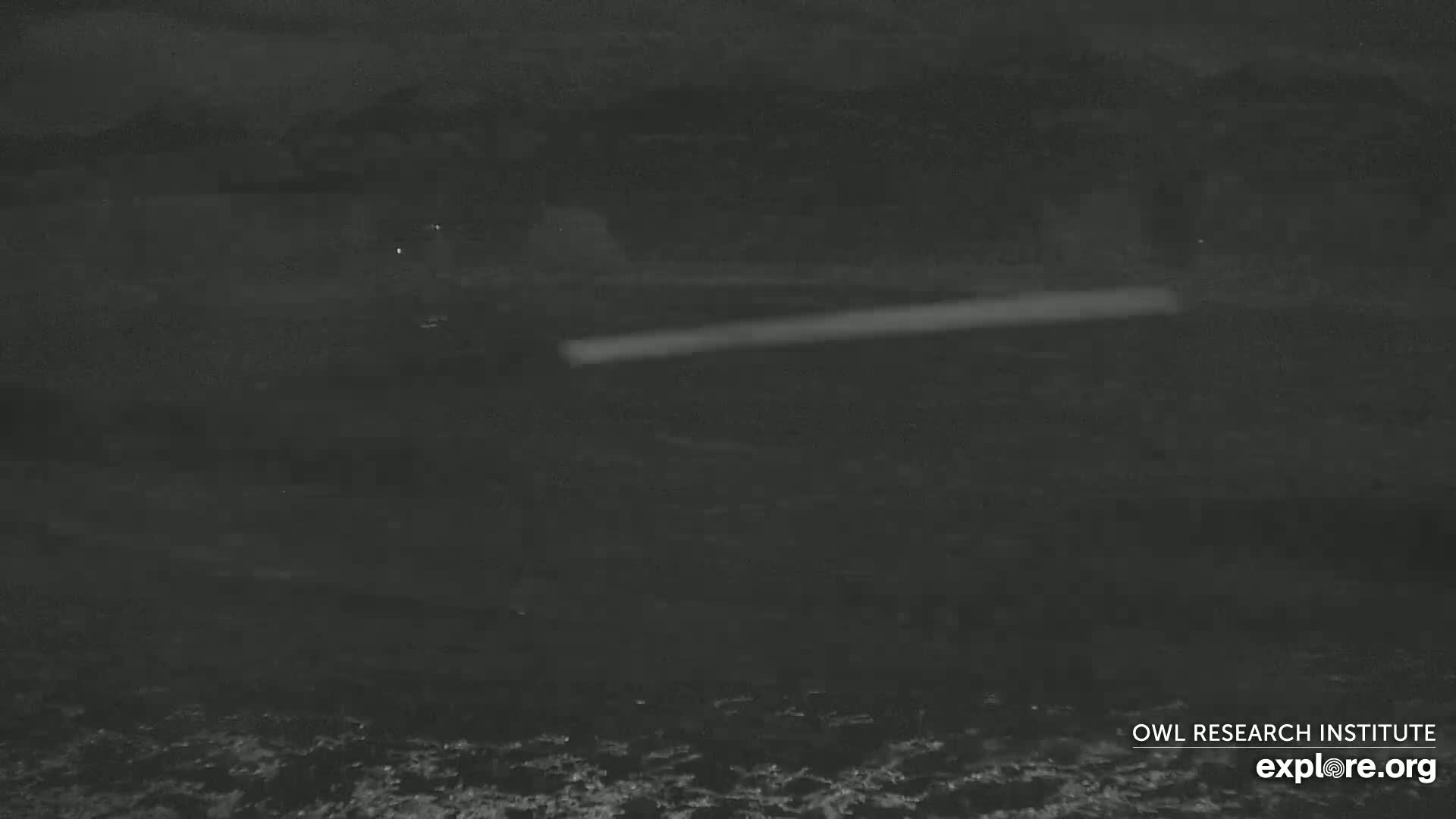 A dark, nighttime scene shows a blurry, bright horizontal line across a dark body of water under a dark sky.