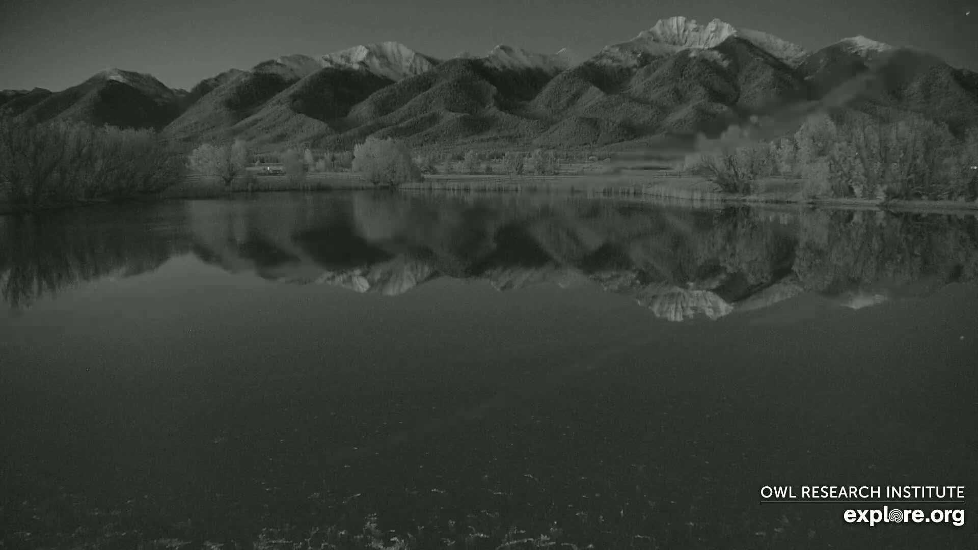 A calm lake reflects a snow-capped mountain range under a dark, clear sky.