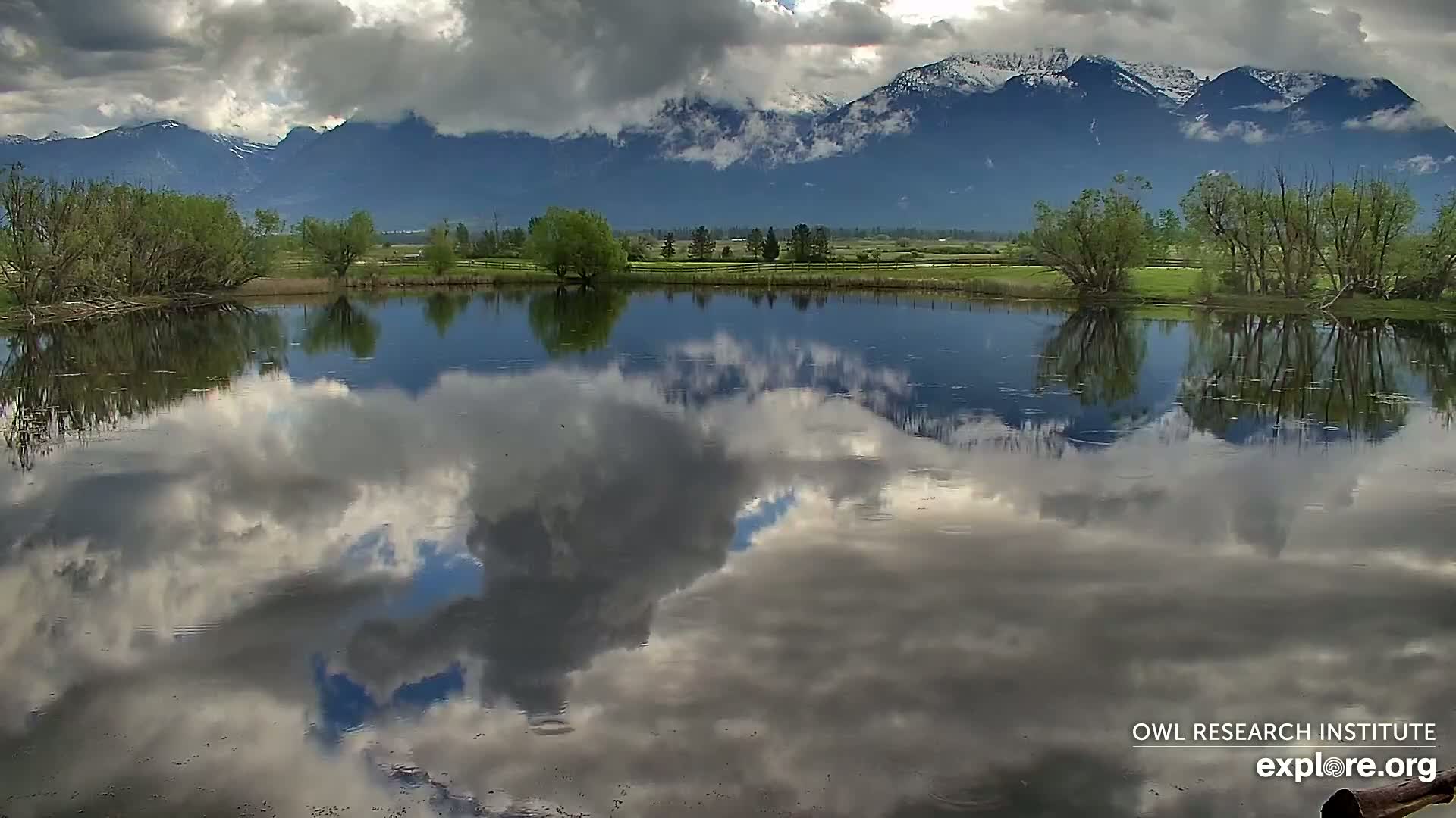A calm pond reflects a cloudy sky and snow-capped mountains under a partly cloudy sky.