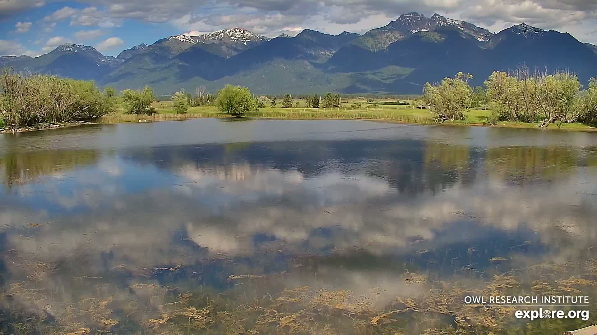 A calm pond reflects a partly cloudy sky and snow-capped mountains under a partly cloudy sky.