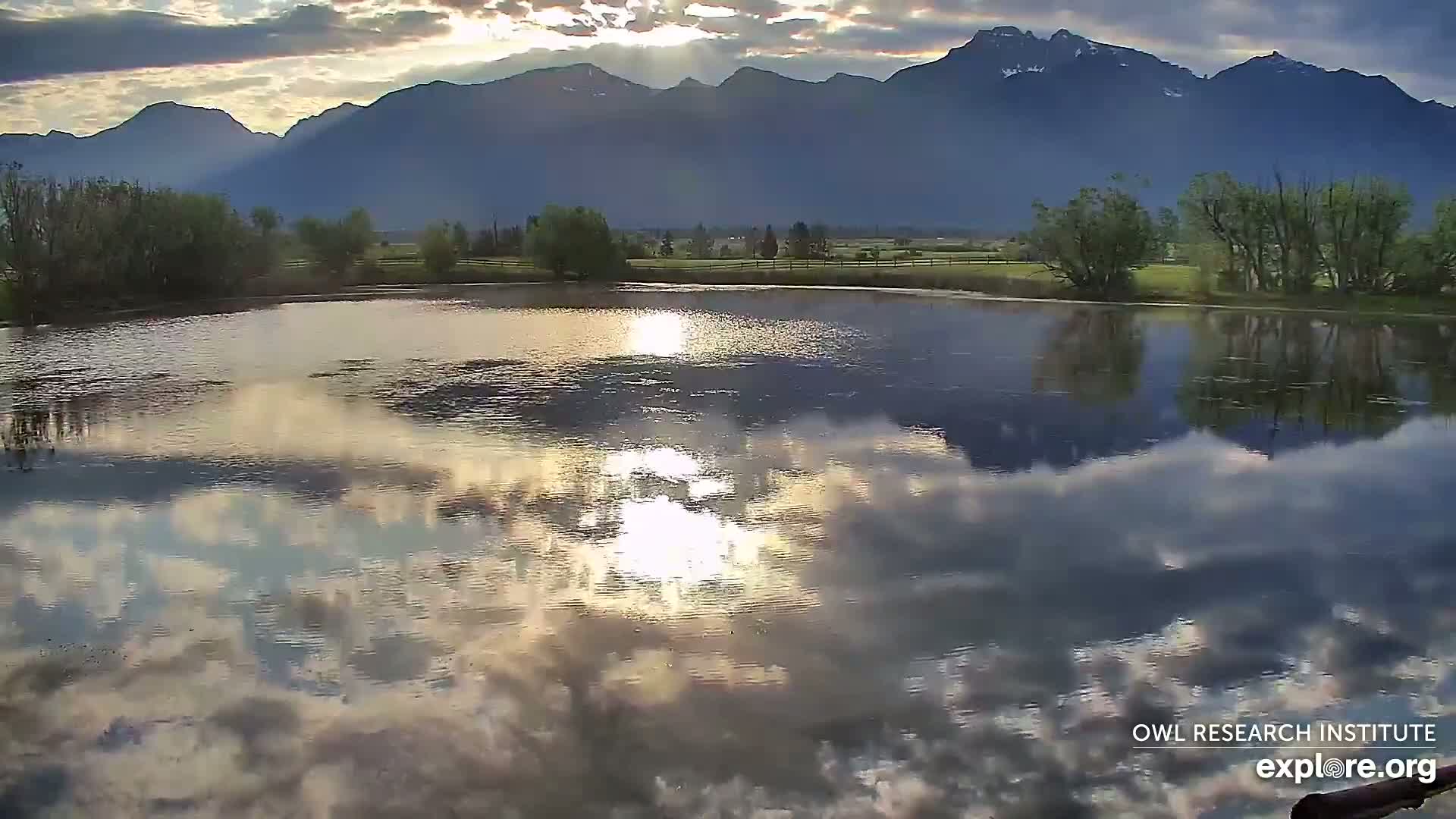 A calm body of water reflects a partly cloudy sky and distant mountains under the soft light of sunrise.