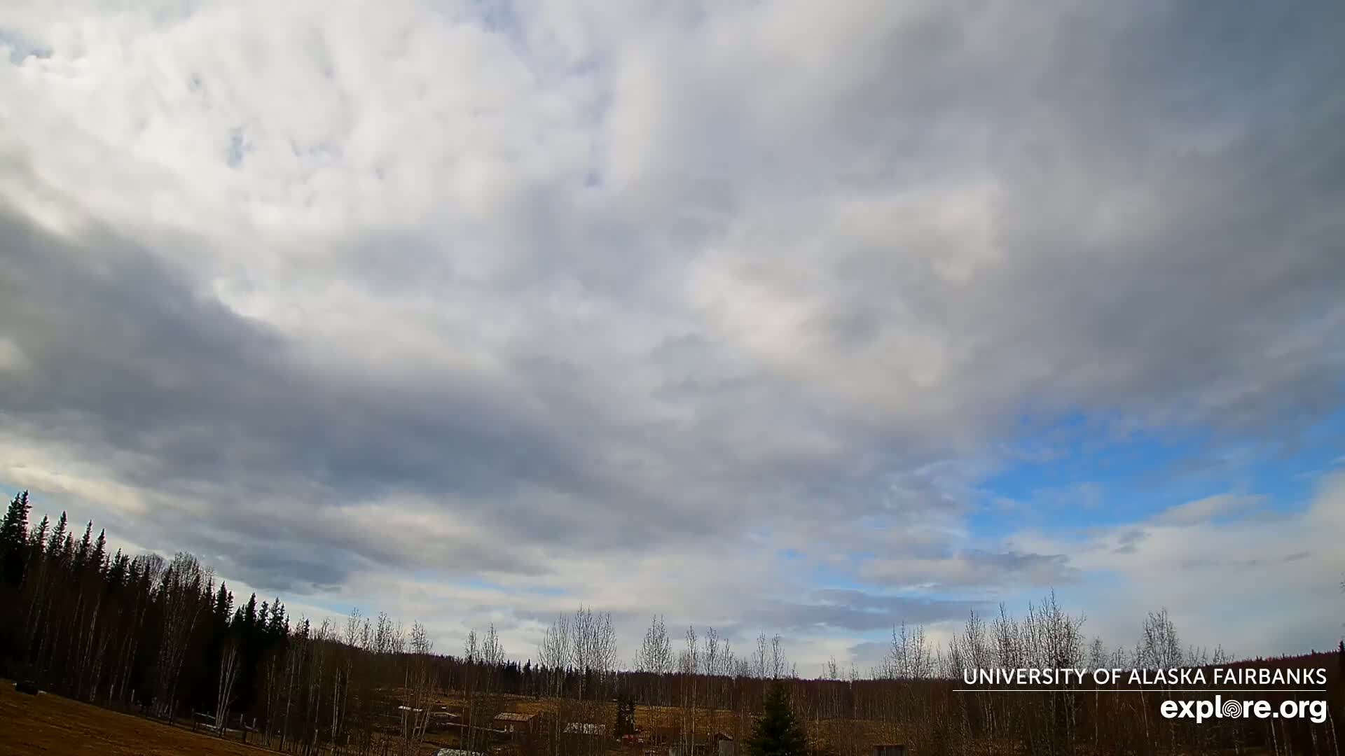 A mostly cloudy sky with patches of blue overhead, above a line of leafless trees and a flat, sparsely developed landscape.