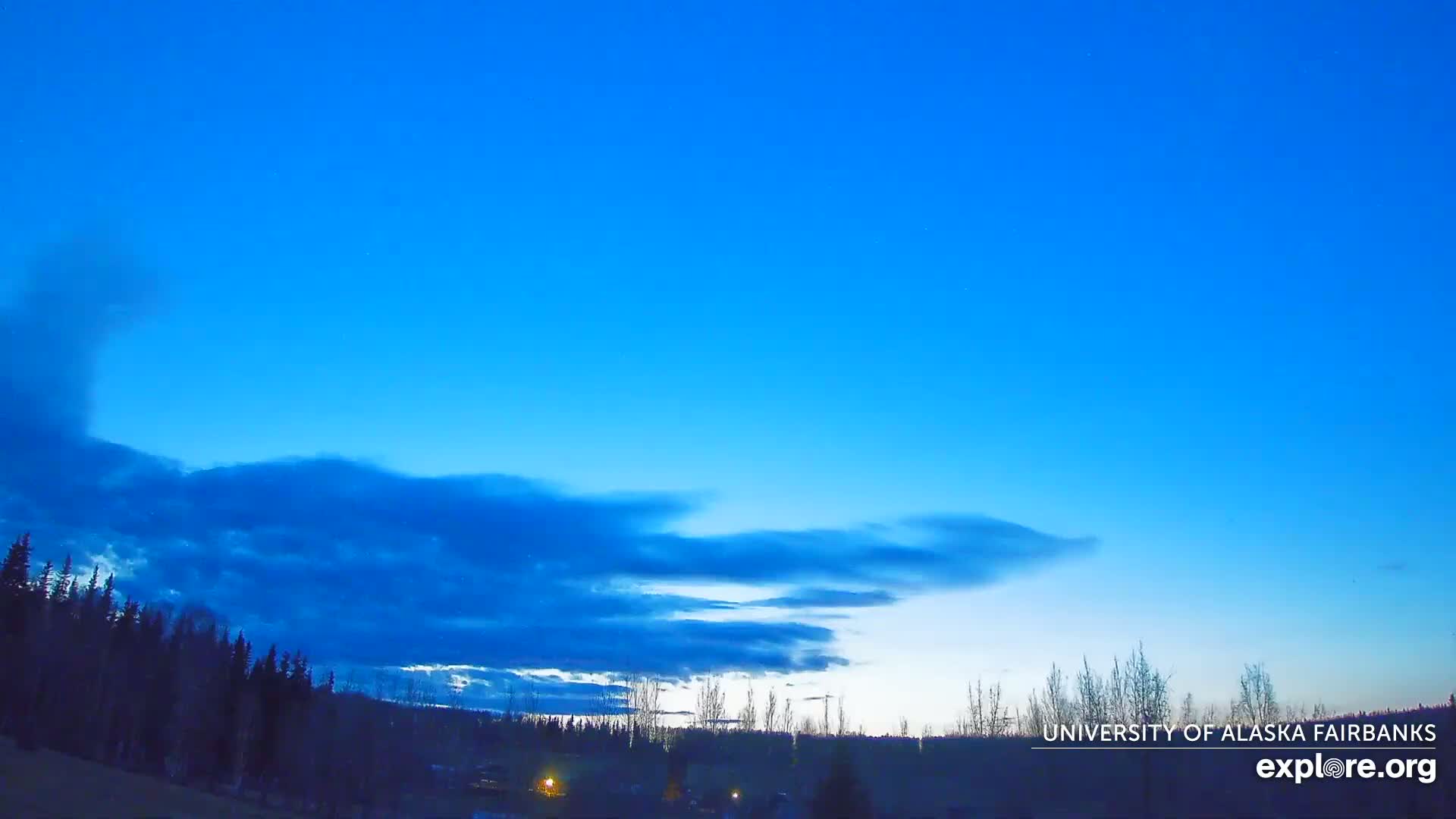 A mostly clear, twilight sky with thin clouds above a dark silhouetted treeline.