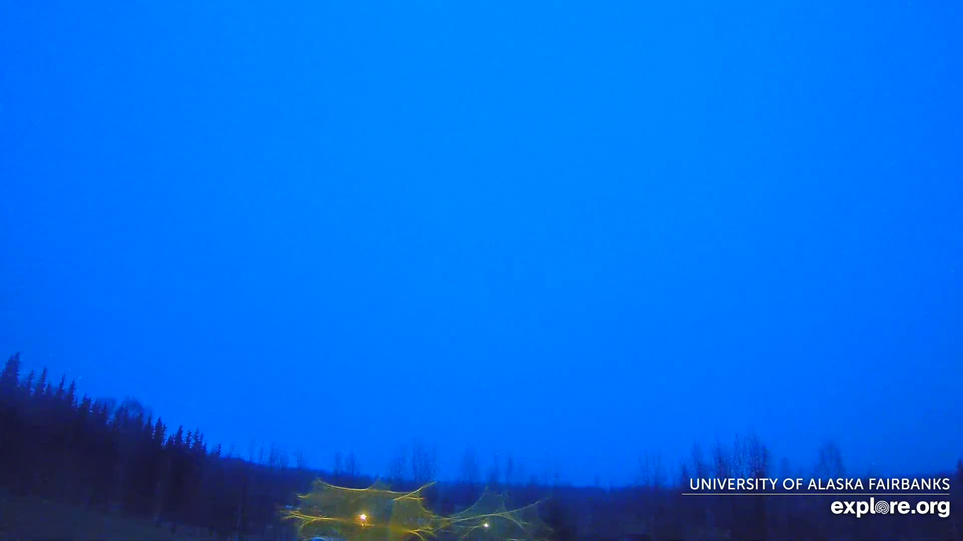 Under a twilight blue sky, a dark line of trees sits at the bottom of the frame, with faint lights visible in the foreground.