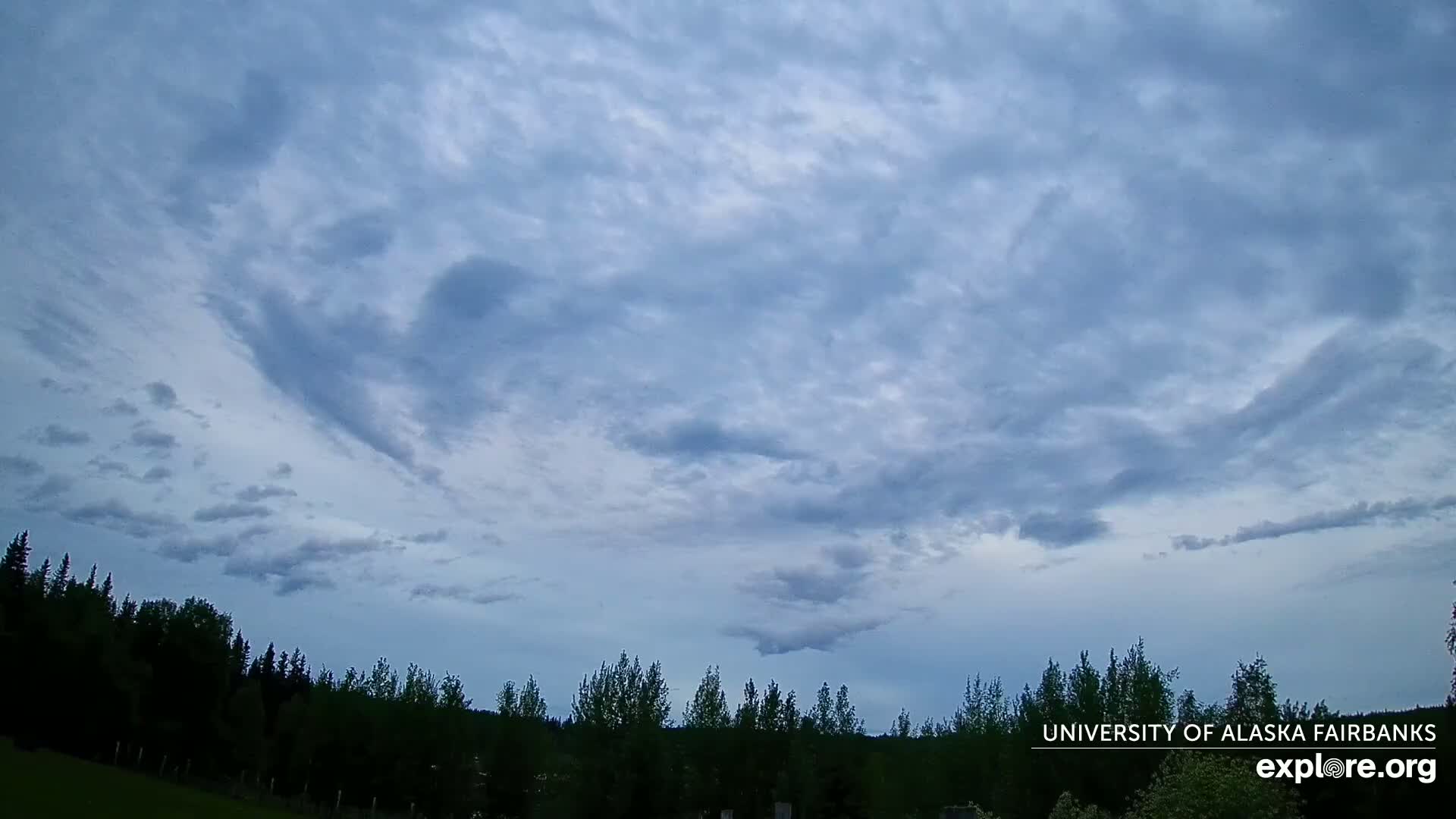 A mostly cloudy sky, appearing somewhat overcast, hangs over a dark silhouetted line of trees.