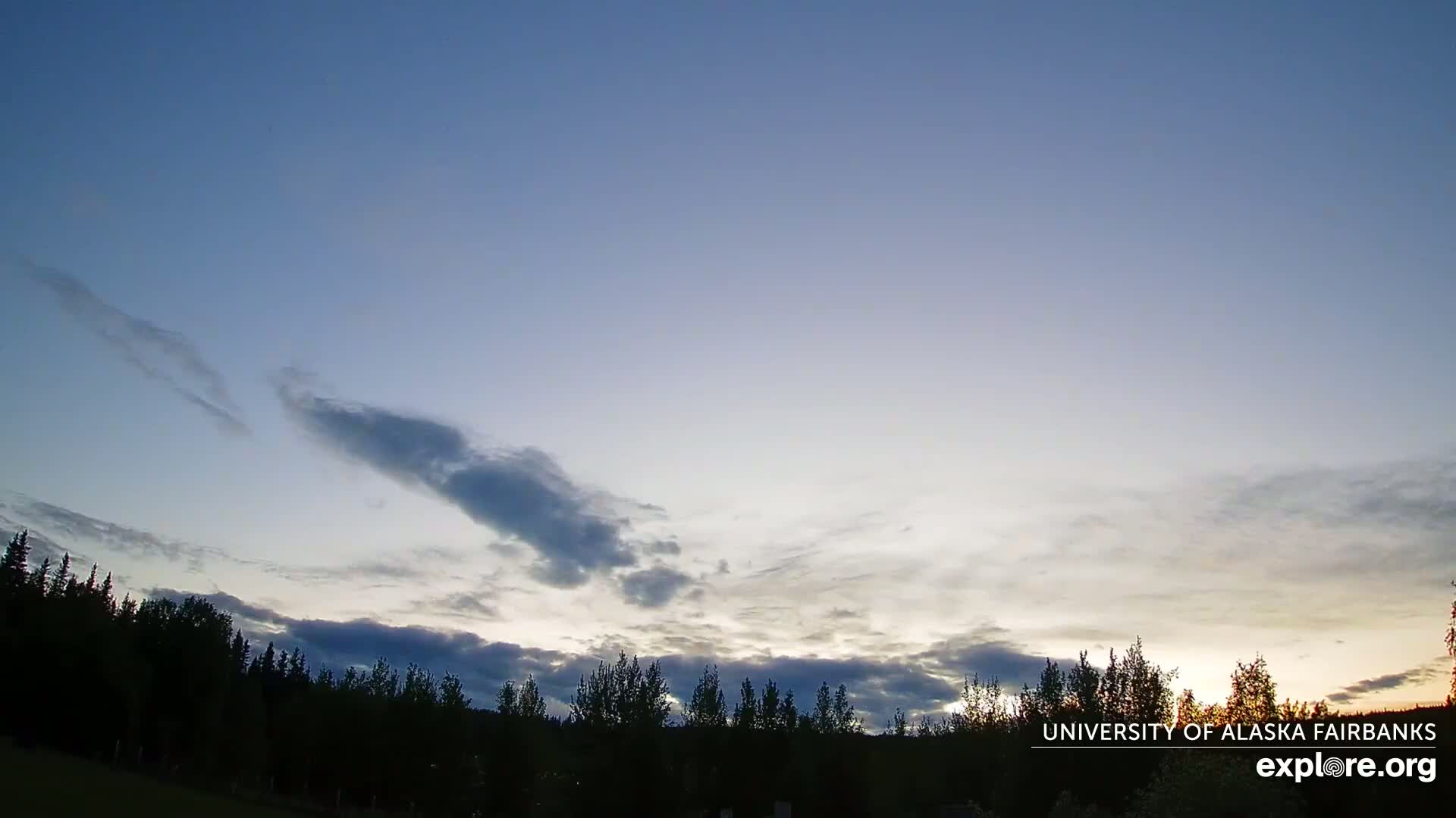 A mostly clear twilight sky with wispy clouds above a dark silhouetted line of trees.