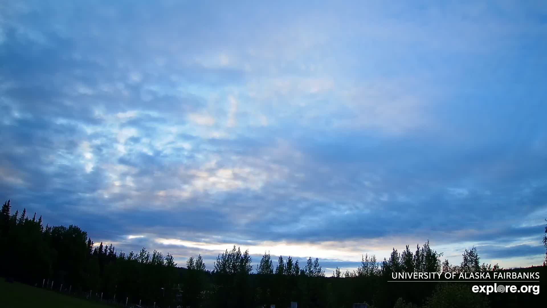 A mostly cloudy, light blue sky at dusk sits above a dark silhouette of a treeline.