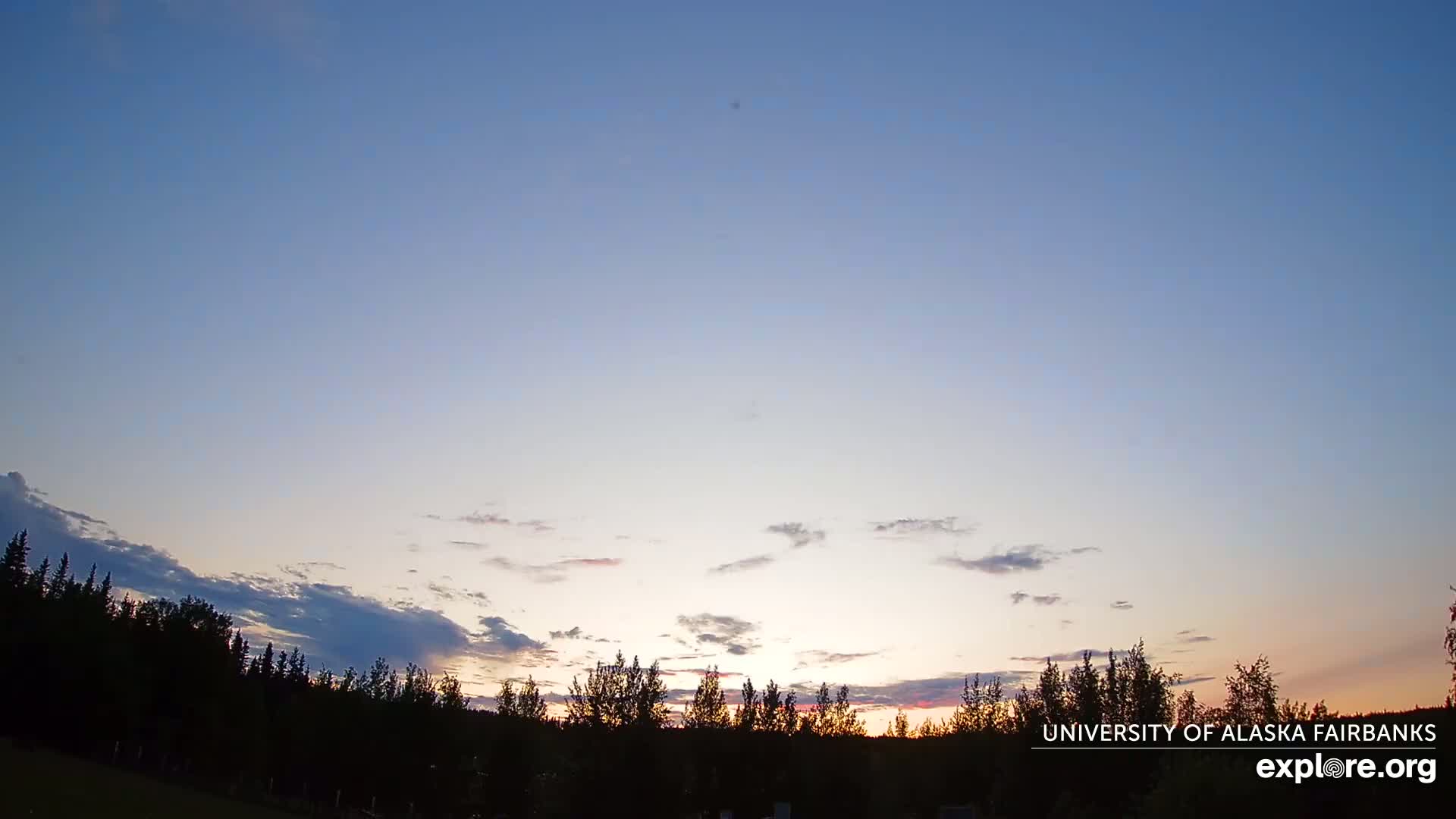 A partly cloudy sunset illuminates a line of silhouetted trees.