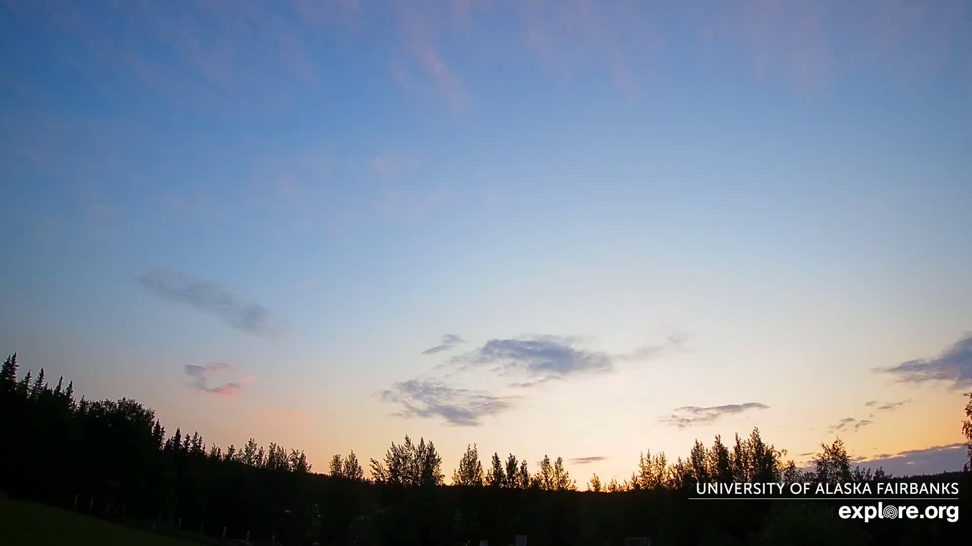 A partly cloudy sunset illuminates a silhouetted line of trees.