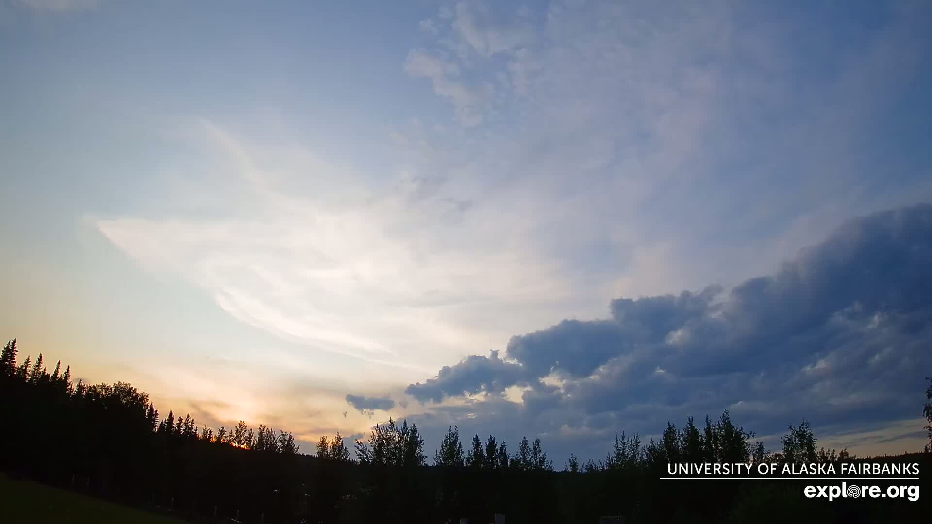 A sunset with partly cloudy skies illuminates a silhouetted line of trees.