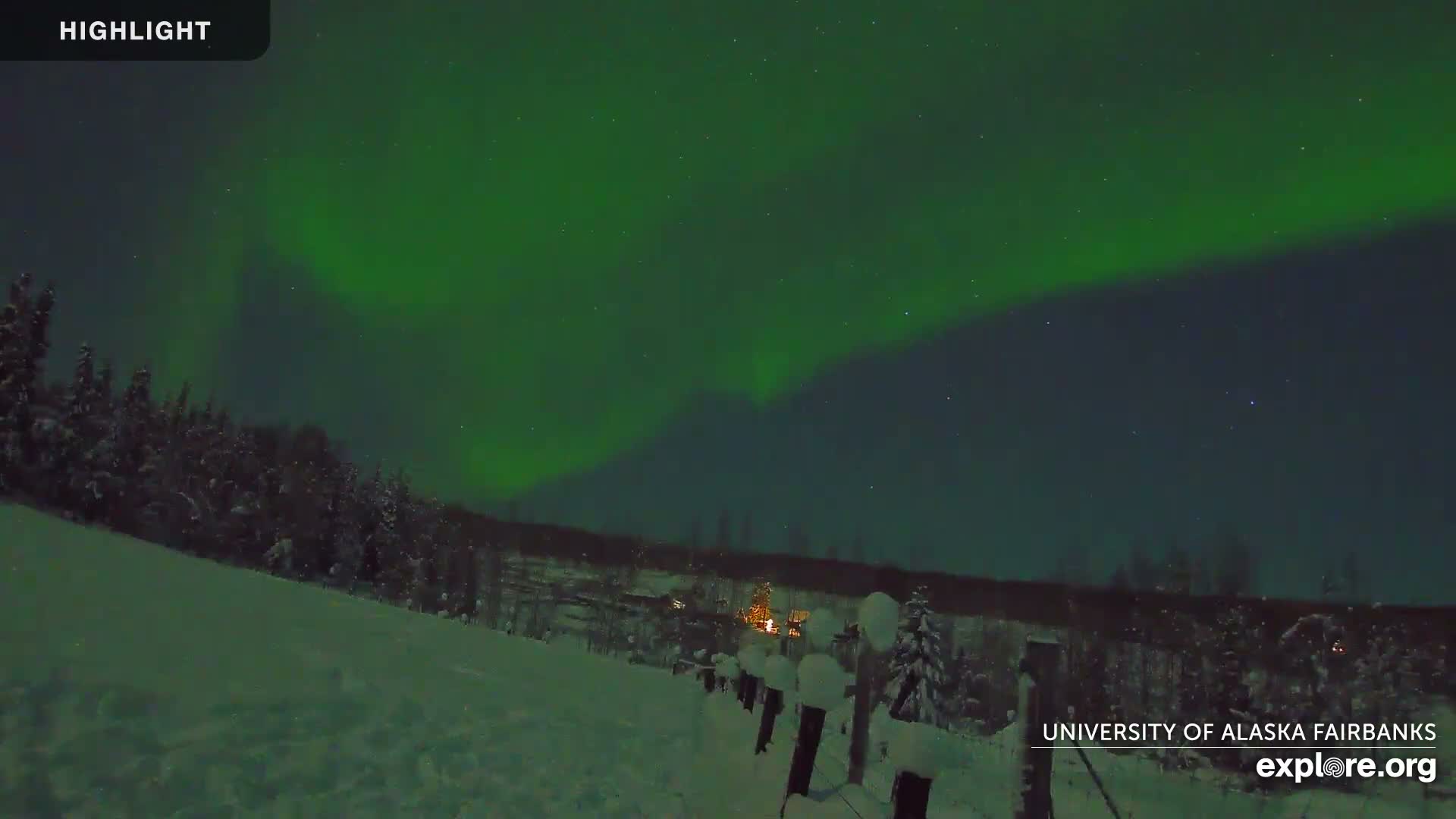 A vibrant green aurora borealis lights up a clear, cold night sky over a snow-covered landscape with distant treelines and a few illuminated structures.