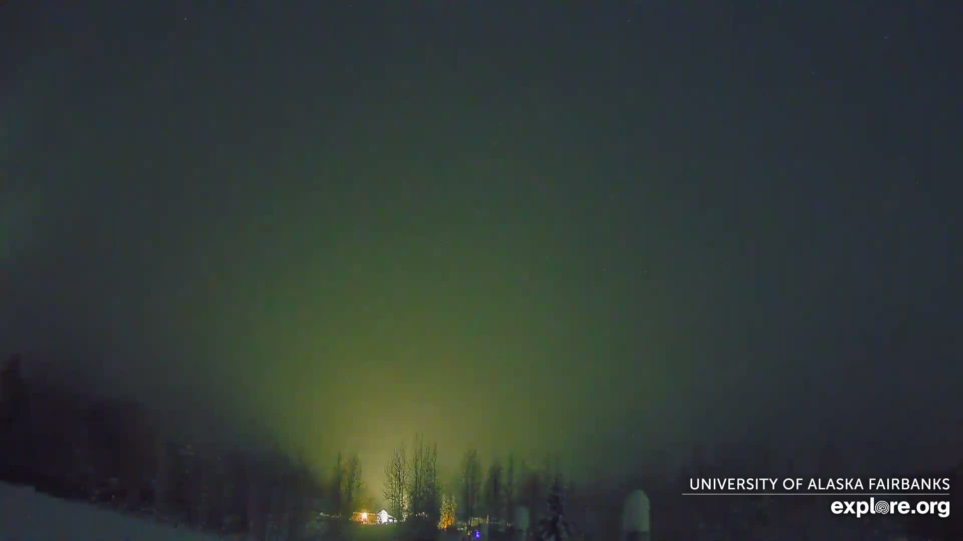 A vibrant green aurora borealis lights up a clear, cold night sky over a snow-covered landscape with distant treelines and a few illuminated structures.