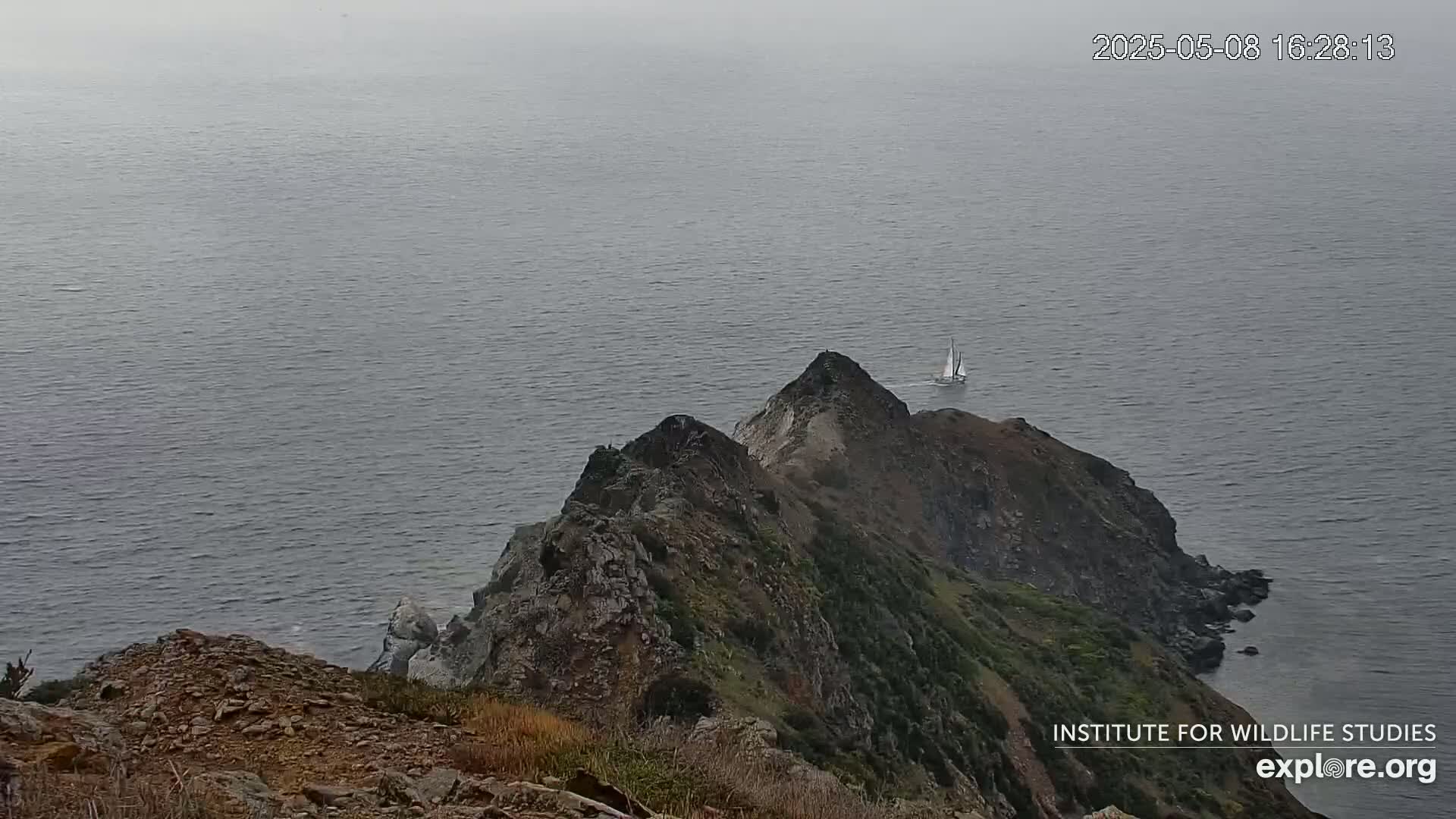 A sailboat is visible on the ocean beyond a rocky, arid coastline under an overcast sky.
