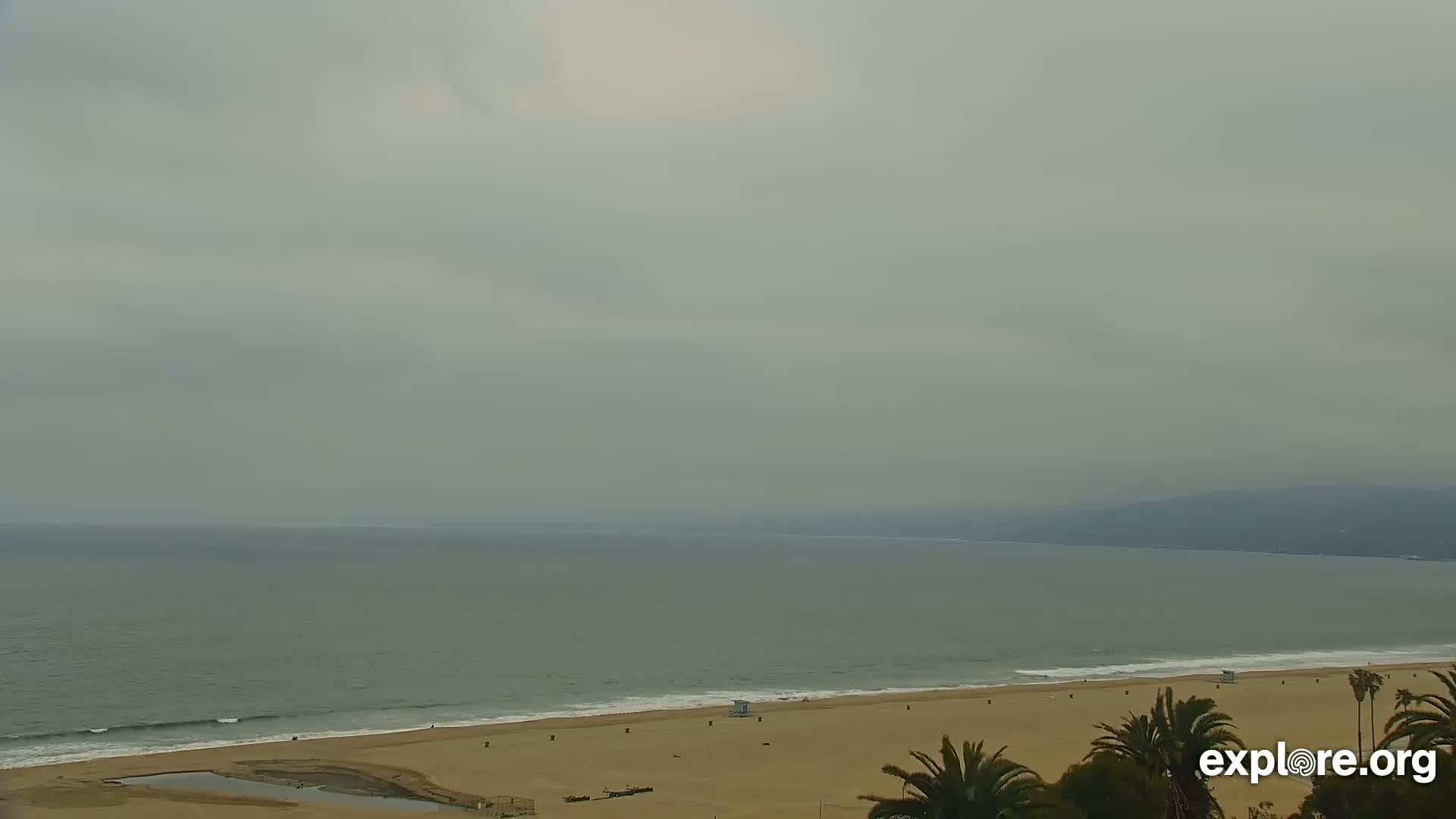 A mostly cloudy day reveals a wide sandy beach with small waves lapping the shore, and a calm ocean extending to a hazy horizon.