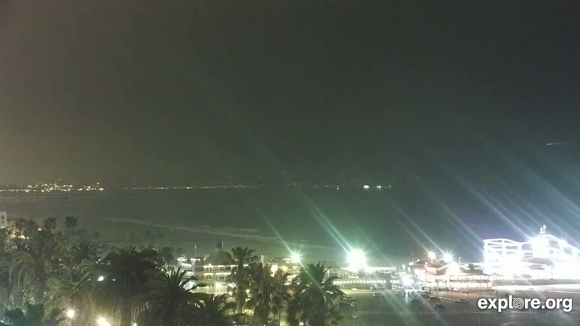 A nighttime view of a beach town with palm trees, a pier, and a body of water, under a hazy sky.