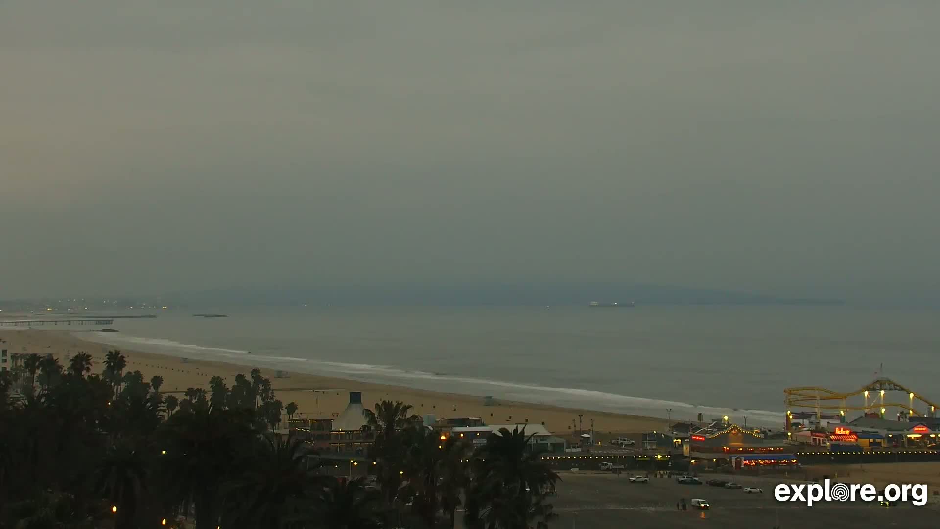 A hazy, overcast day reveals a long sandy beach with a pier and amusement park, and palm trees lining the shore.