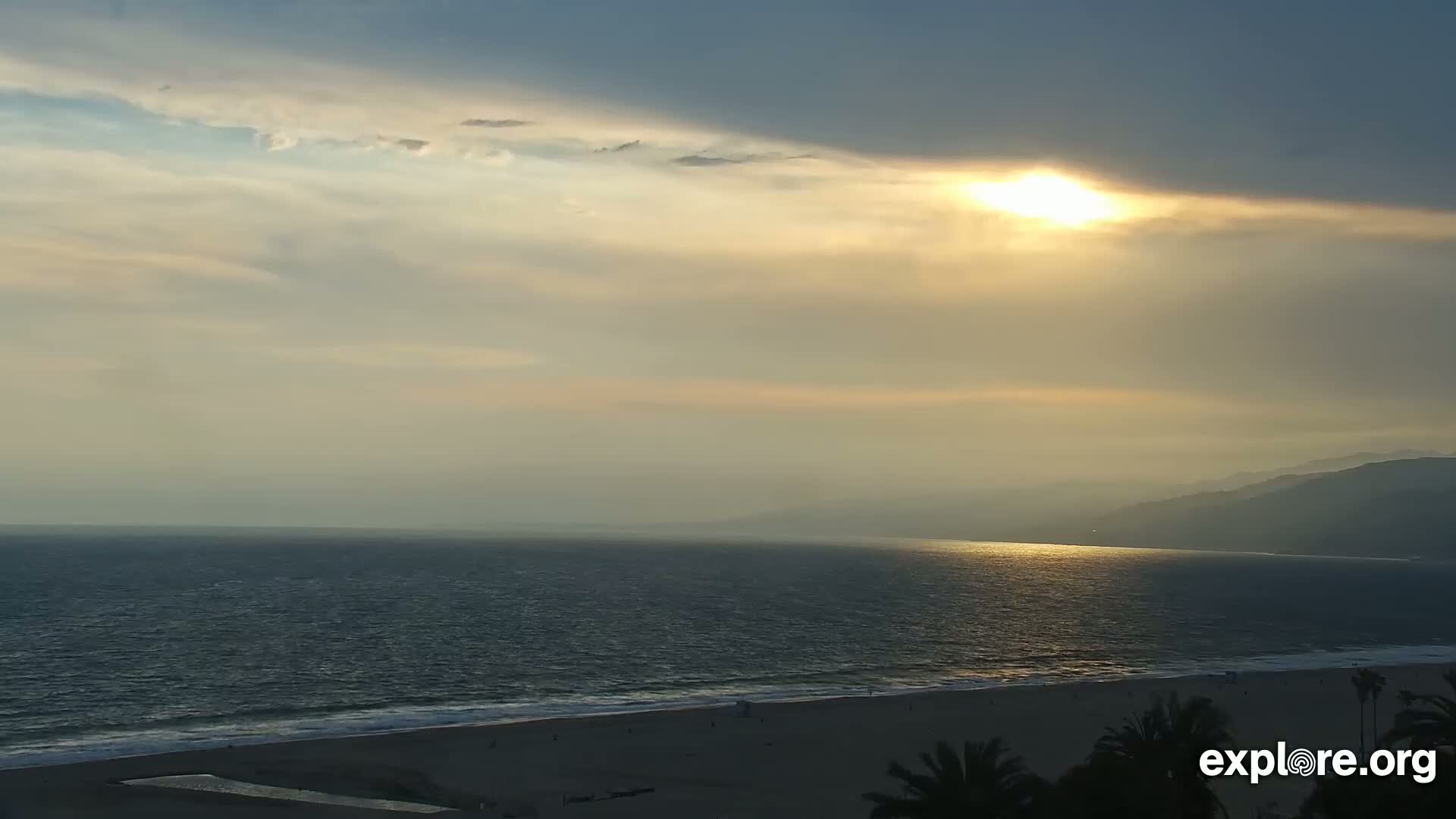 A hazy sunset over a calm ocean and sandy beach, with distant mountains visible on the horizon.
