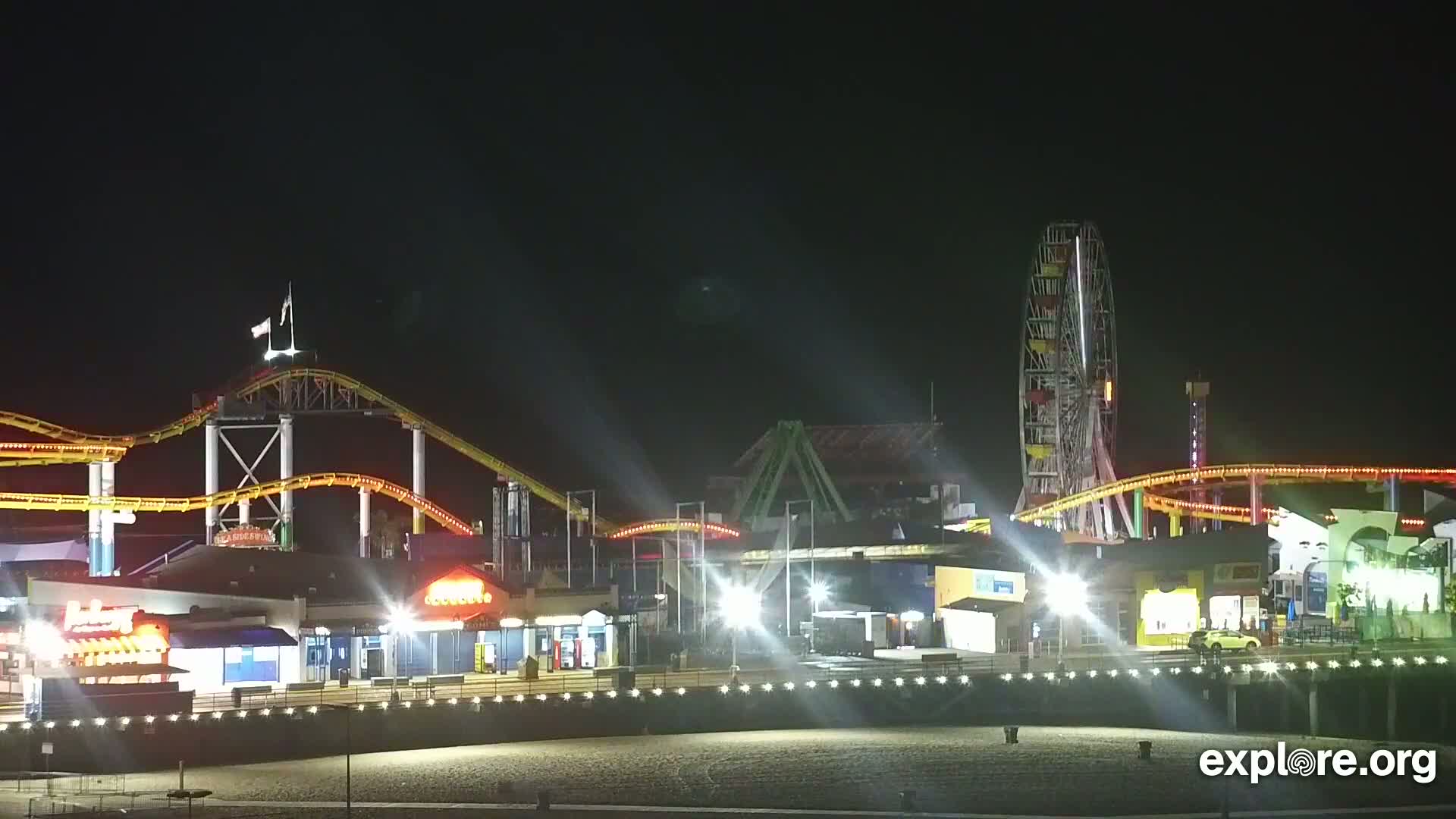 At night, brightly lit rollercoasters and a Ferris wheel are visible on a pier overlooking a sandy beach.